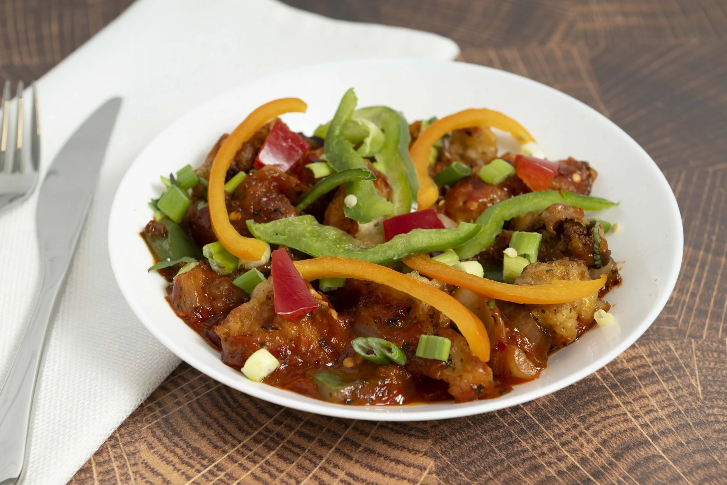 A white bowl filled with stew containing pieces of meat, red and green bell peppers, and chopped green onions, on a wooden table with a fork and knife on a napkin nearby.
