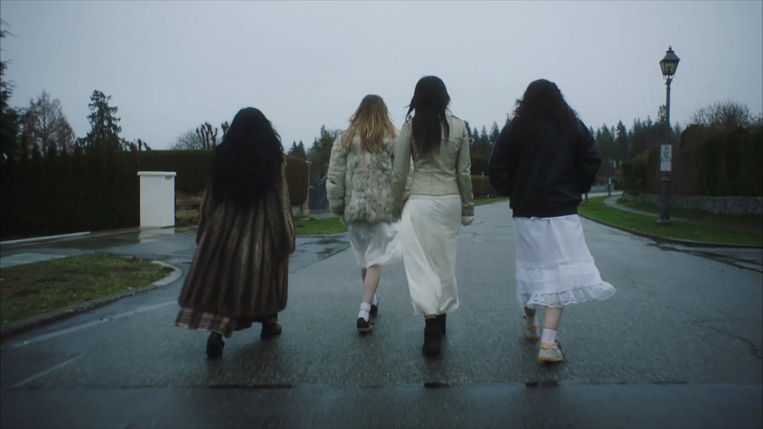 Four women dressed in vintage clothing walking on a wet road in a rural or suburban neighborhood under gray, overcast sky.