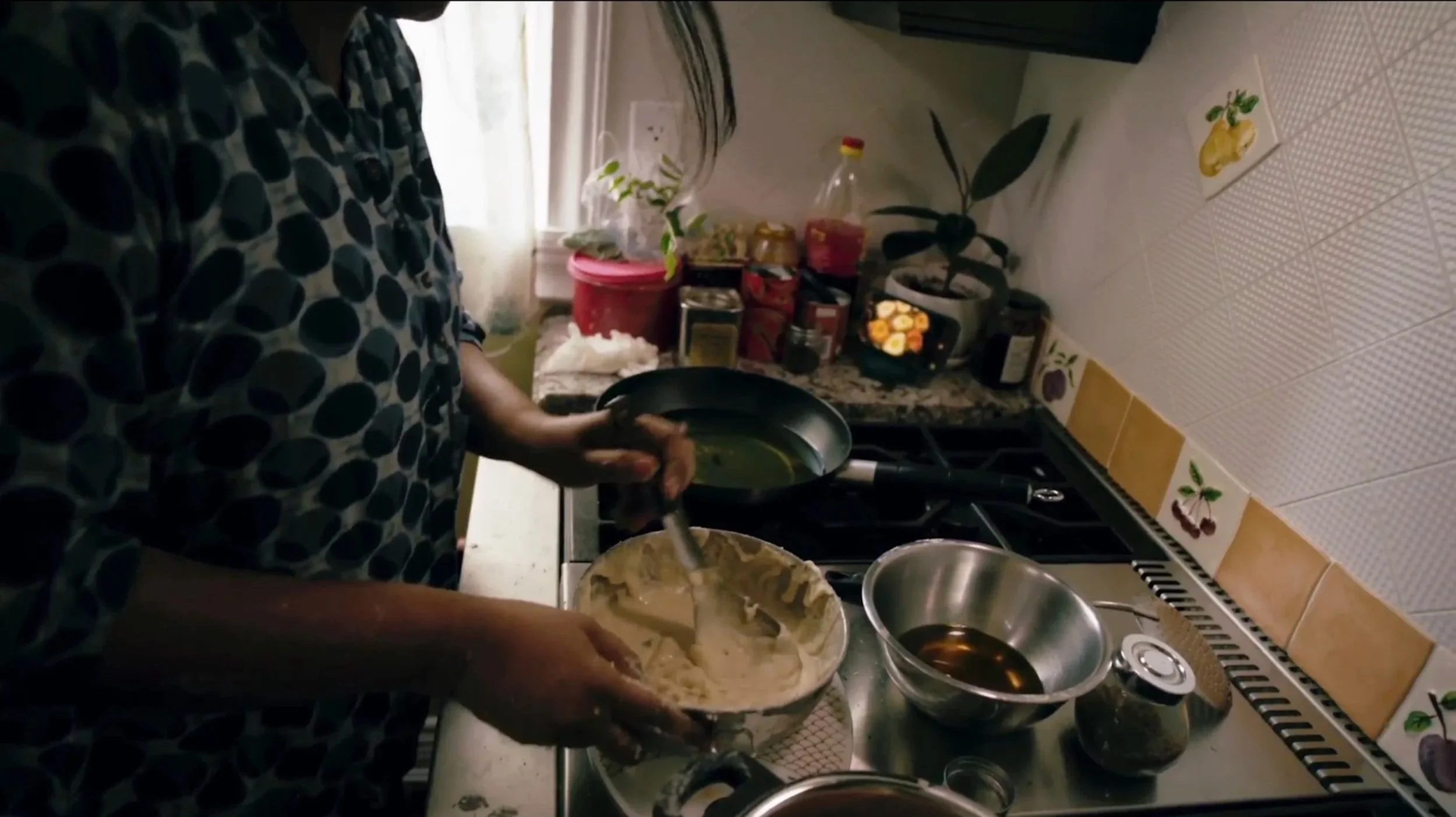 Person cooking in a kitchen, holding a black frying pan, with bowls and food on the countertop, and various food items and jars in the background.