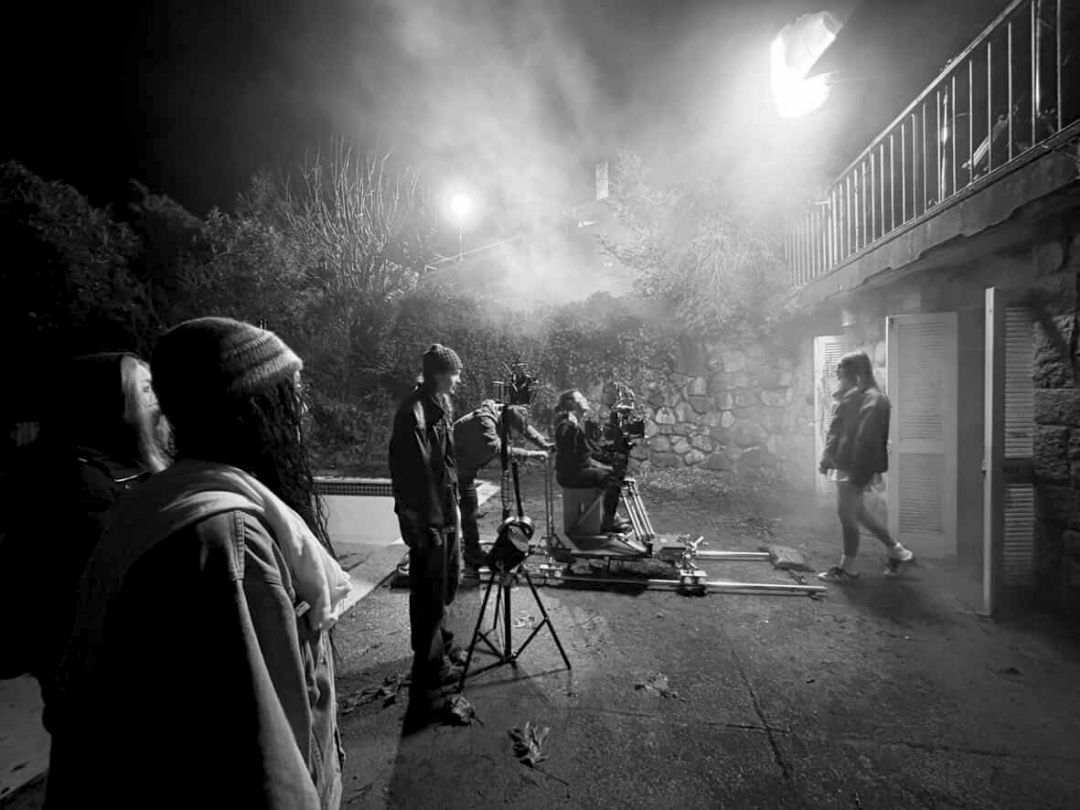 Film crew working outdoors at night, filming a scene with a person walking out of a building, with lights and smoke in the background.