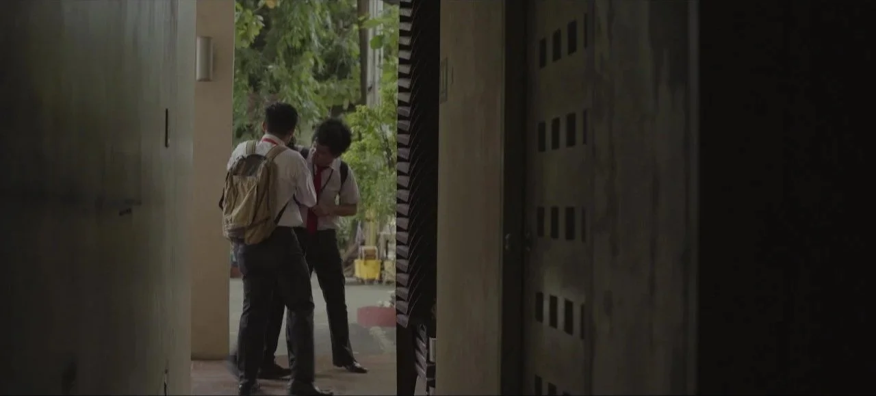 Two students in school uniforms and backpacks standing on a sidewalk outside, talking to each other near a building entrance, with lush green trees in the background.