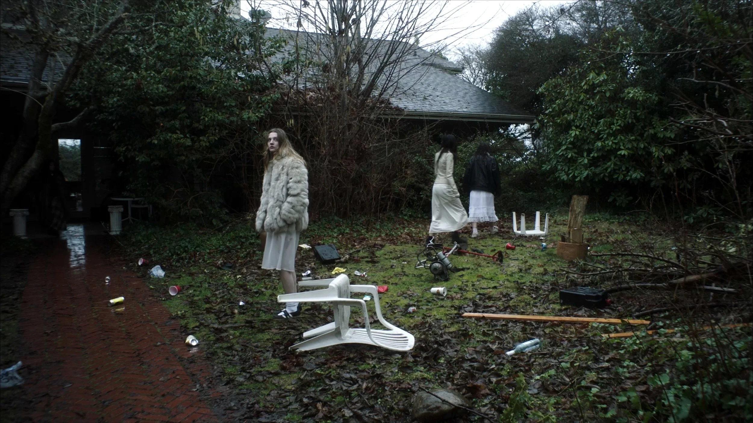 Disorded backyard with trash and debris, three women standing among broken furniture and scattered items, surrounded by trees and bushes.