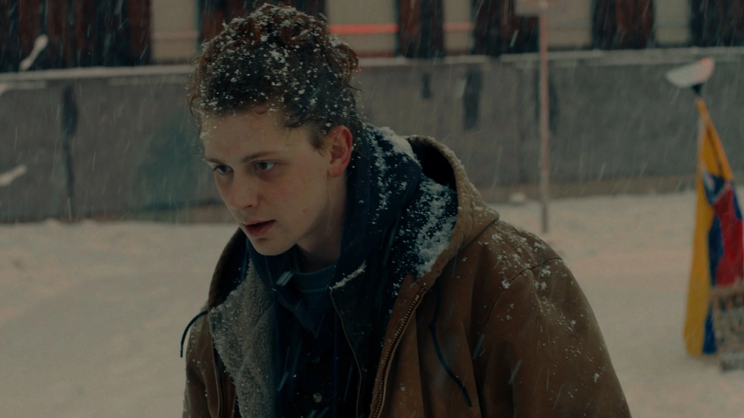 A young man with curly hair leaning forward outdoors in snowy weather, wearing a brown jacket with a shearling collar and a dark hoodie. Snow covers his hair and shoulders, and a blurred backdrop of a fence and a colorful flag is visible.