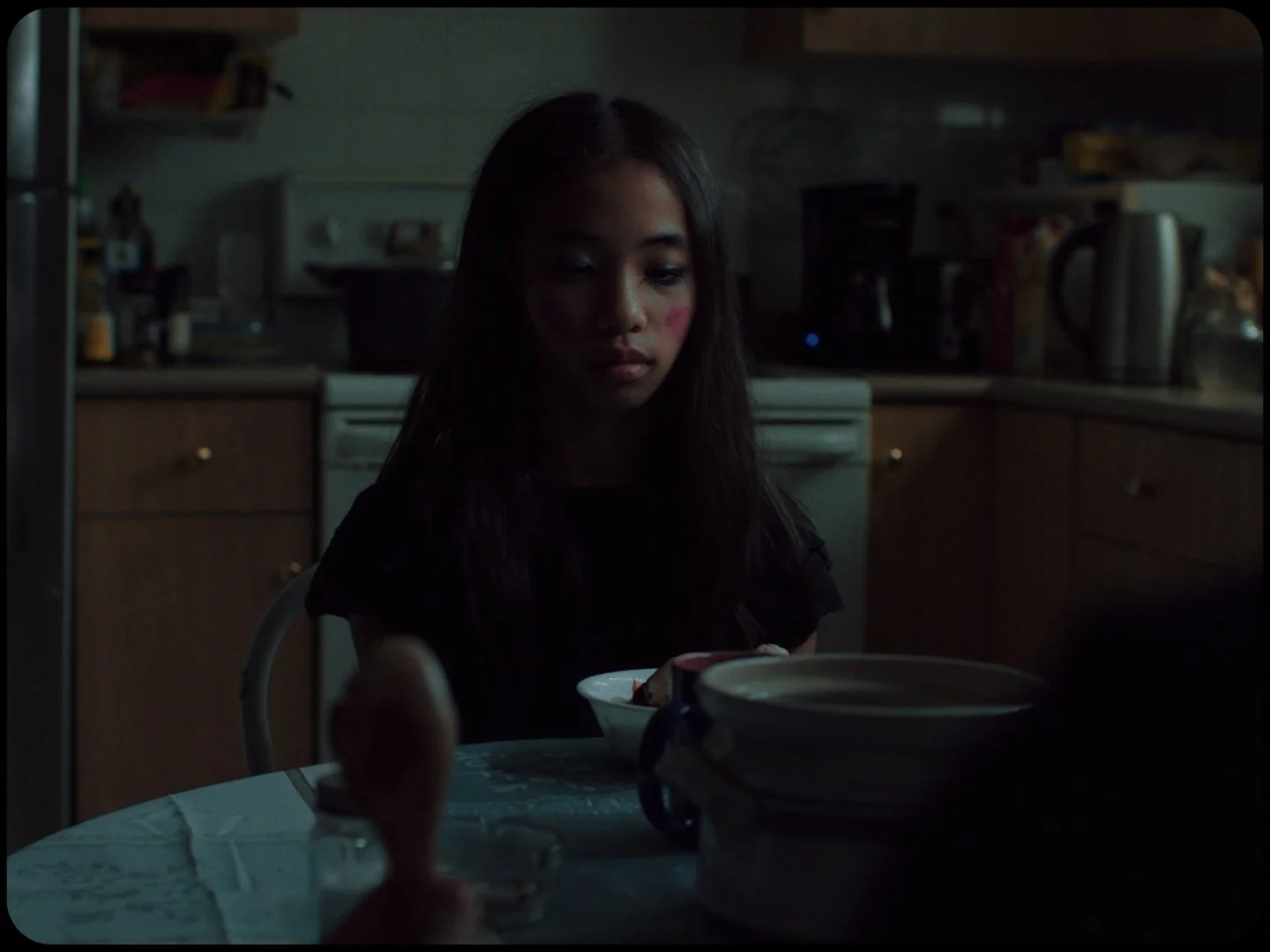 A young woman with long dark hair sitting at a kitchen table in a dimly lit kitchen, with a bowl in front of her and a coffee cup nearby.
