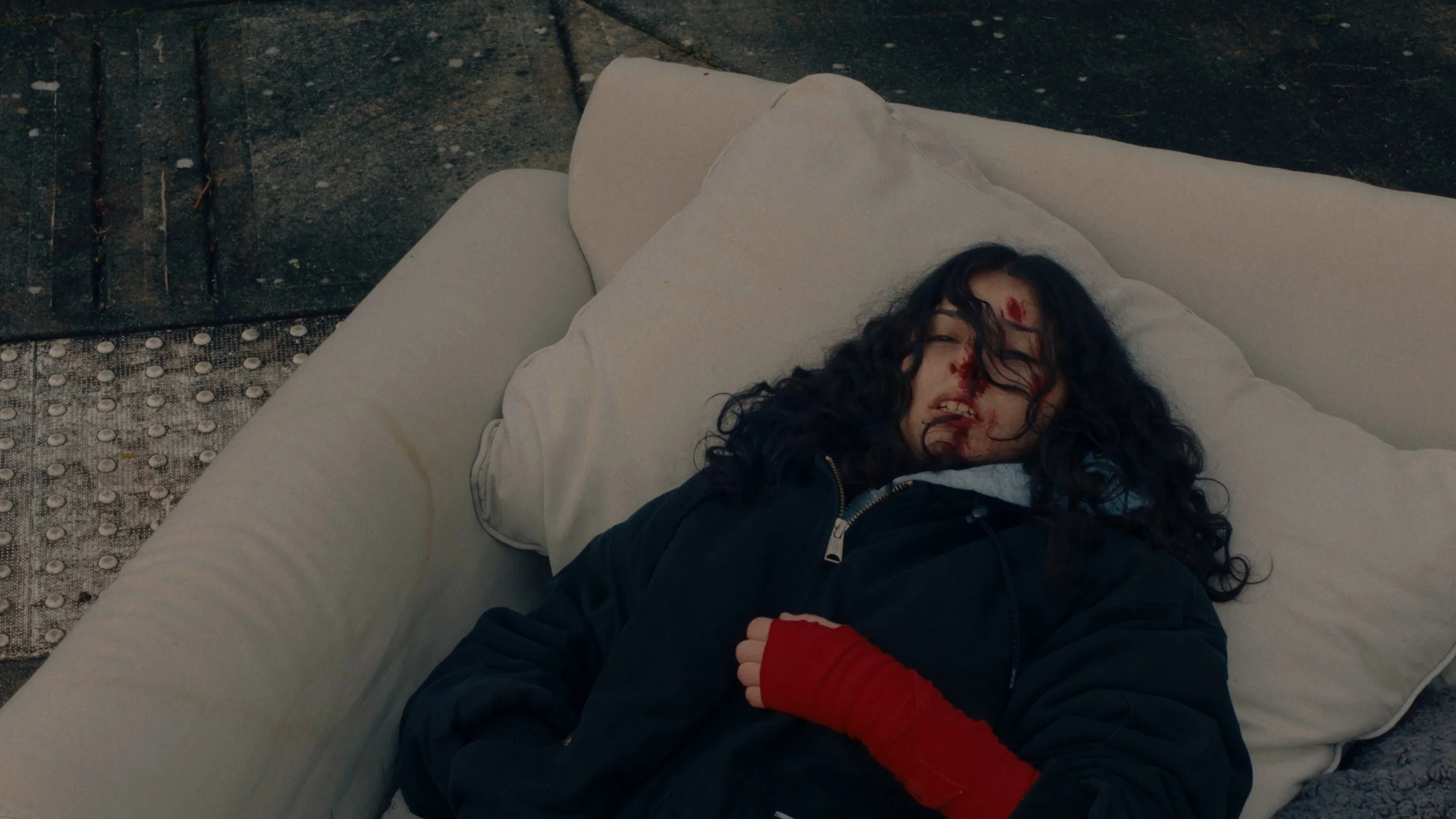 A woman lying on her back on a white couch with blood on her forehead and face, wearing a black jacket and red gloves.