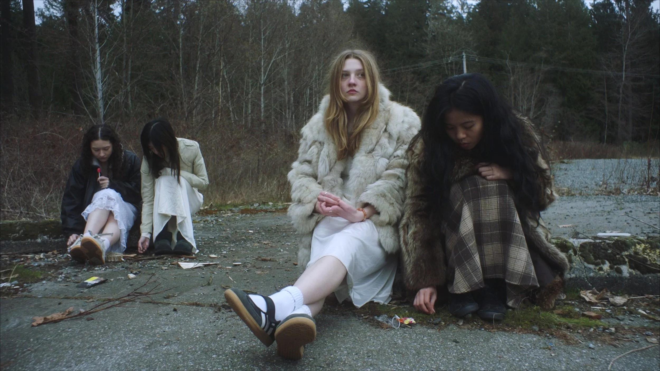 Four young women sitting on the ground outdoors in a natural setting with trees in the background. Two women in the foreground are dressed in vintage or fancy clothing, one in a white dress with a faux fur coat and the other in a plaid coat with a fur collar. The two women in the background are dressed in modern casual clothing, one with a white dress and the other with a black jacket, looking down at the ground.