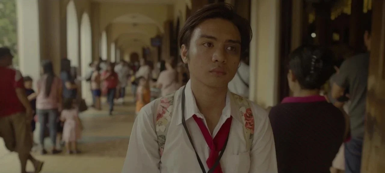 A young man in a white shirt with a red tie, carrying a backpack, standing in a crowded, arched corridor or walkway, with people and children in the background.