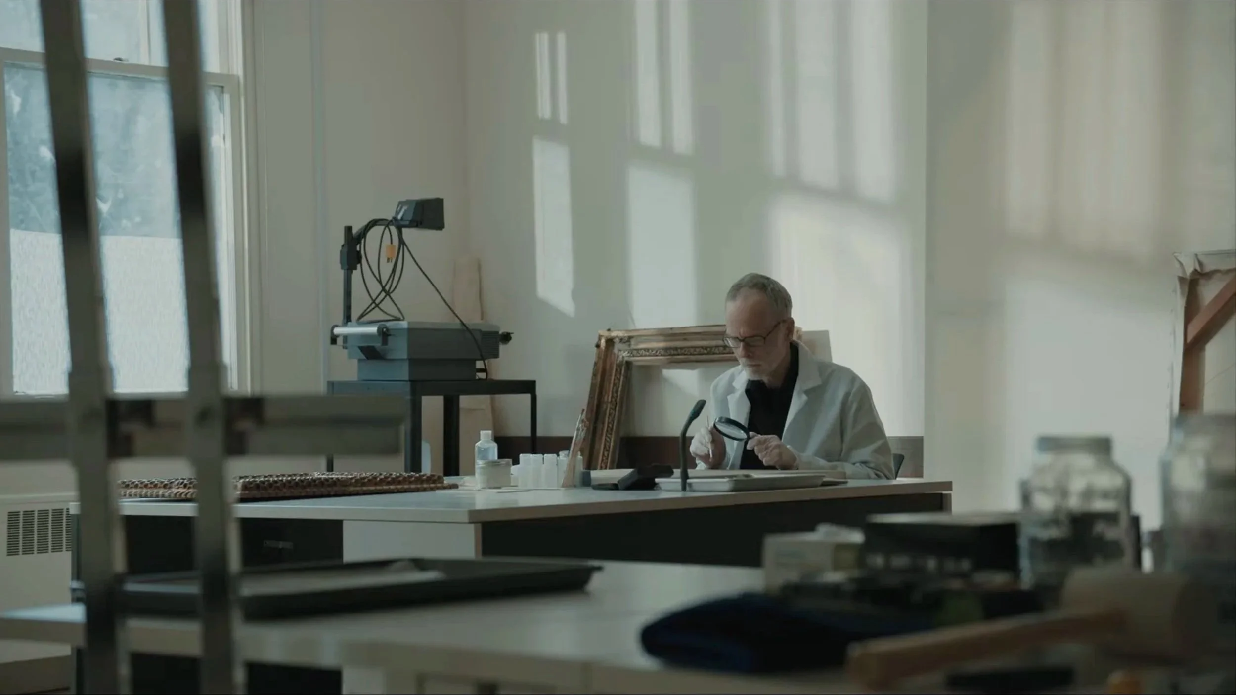 A man in a white lab coat sitting at a table in a well-lit room, examining an object with a magnifying glass.