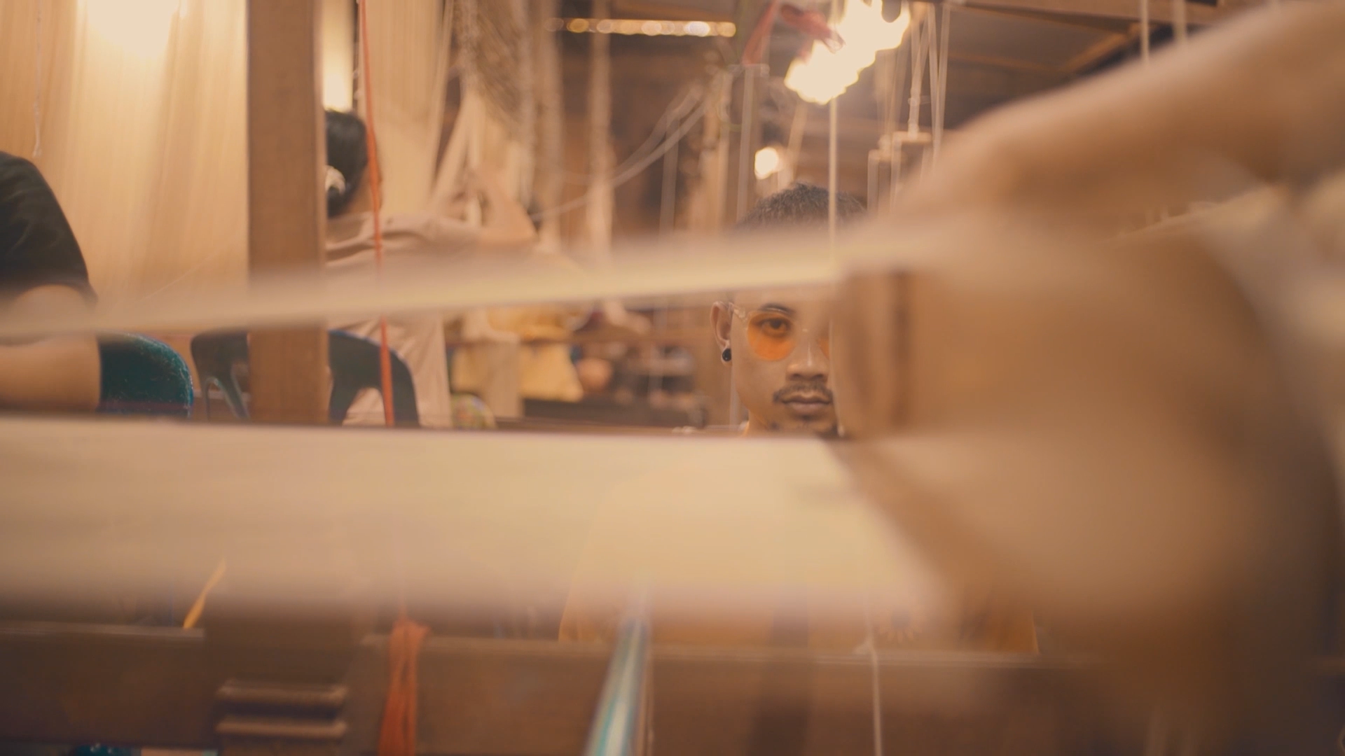 A person wearing orange-tinted glasses and earrings looking through a mirror in a cluttered workshop or studio, with various equipment and materials around.
