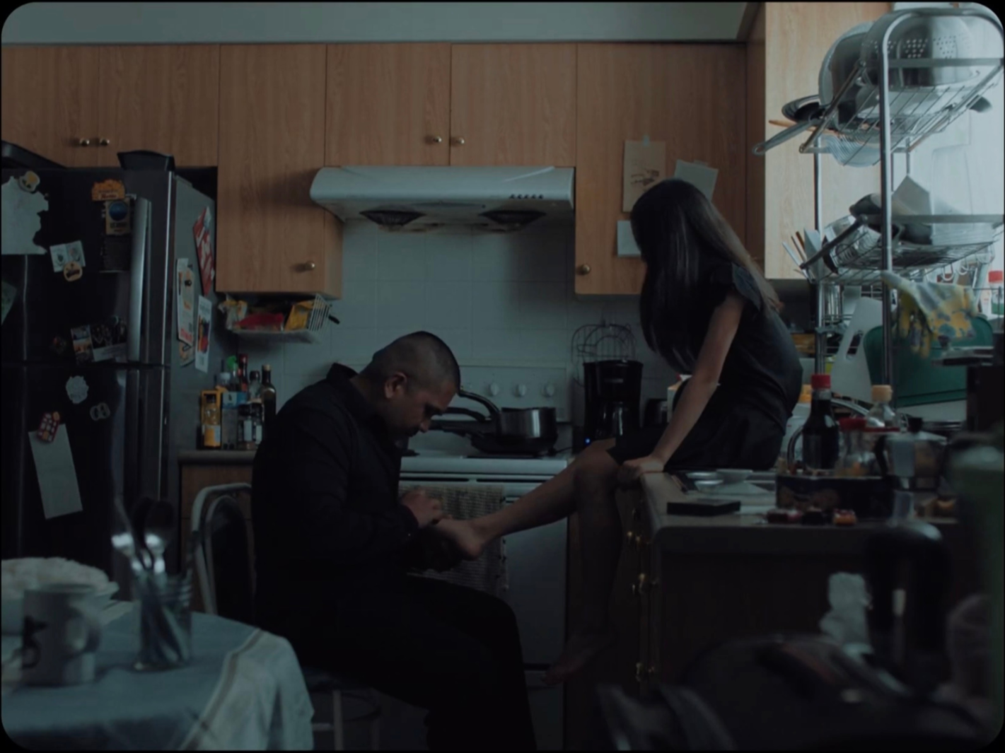A man and a woman in a cluttered kitchen, the man sitting and holding the woman's foot, while the woman leans on the counter, both engaged in an intimate moment.