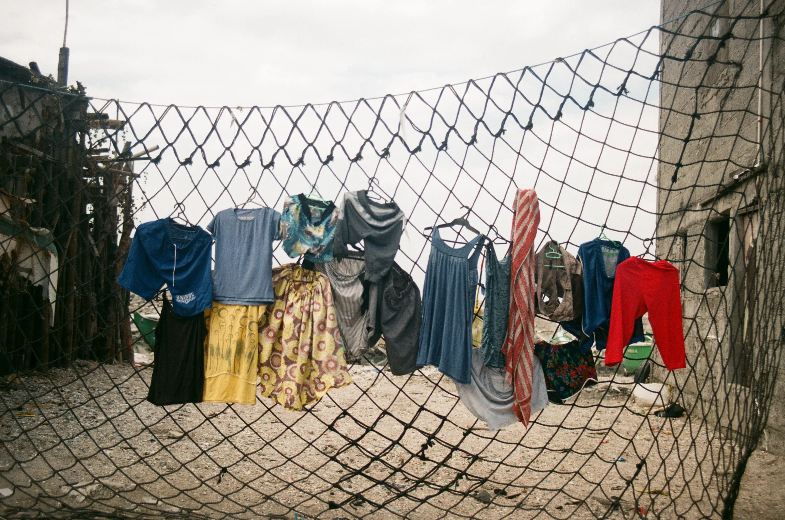 Clothes hanging on a wire fence in an outdoor area with debris and worn buildings.
