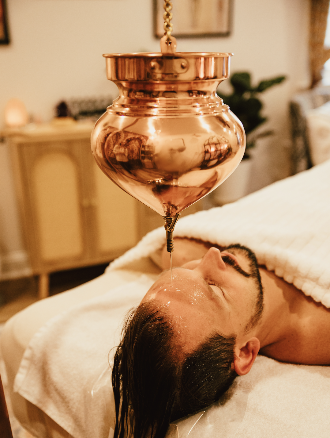 A man lying on a massage table receiving an Ayurvedic Shirodhara treatment with a copper soaking jar suspended above his forehead while warm medicated oil is continuously pored during a holistic therapy session in a cozy, warmly lit room.