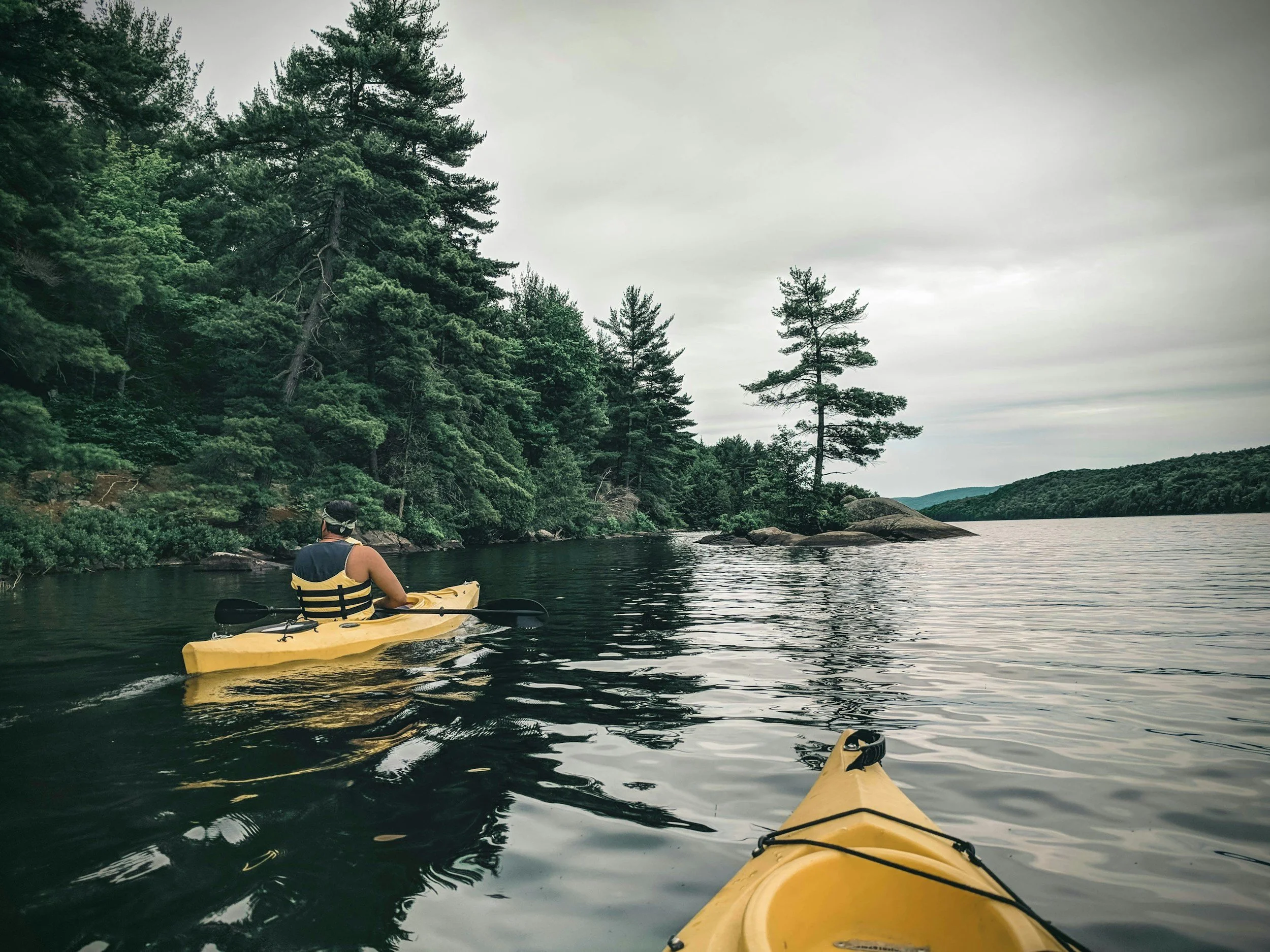 Person kayaking on a calm lake surrounded by green trees and rocks under a cloudy sky.