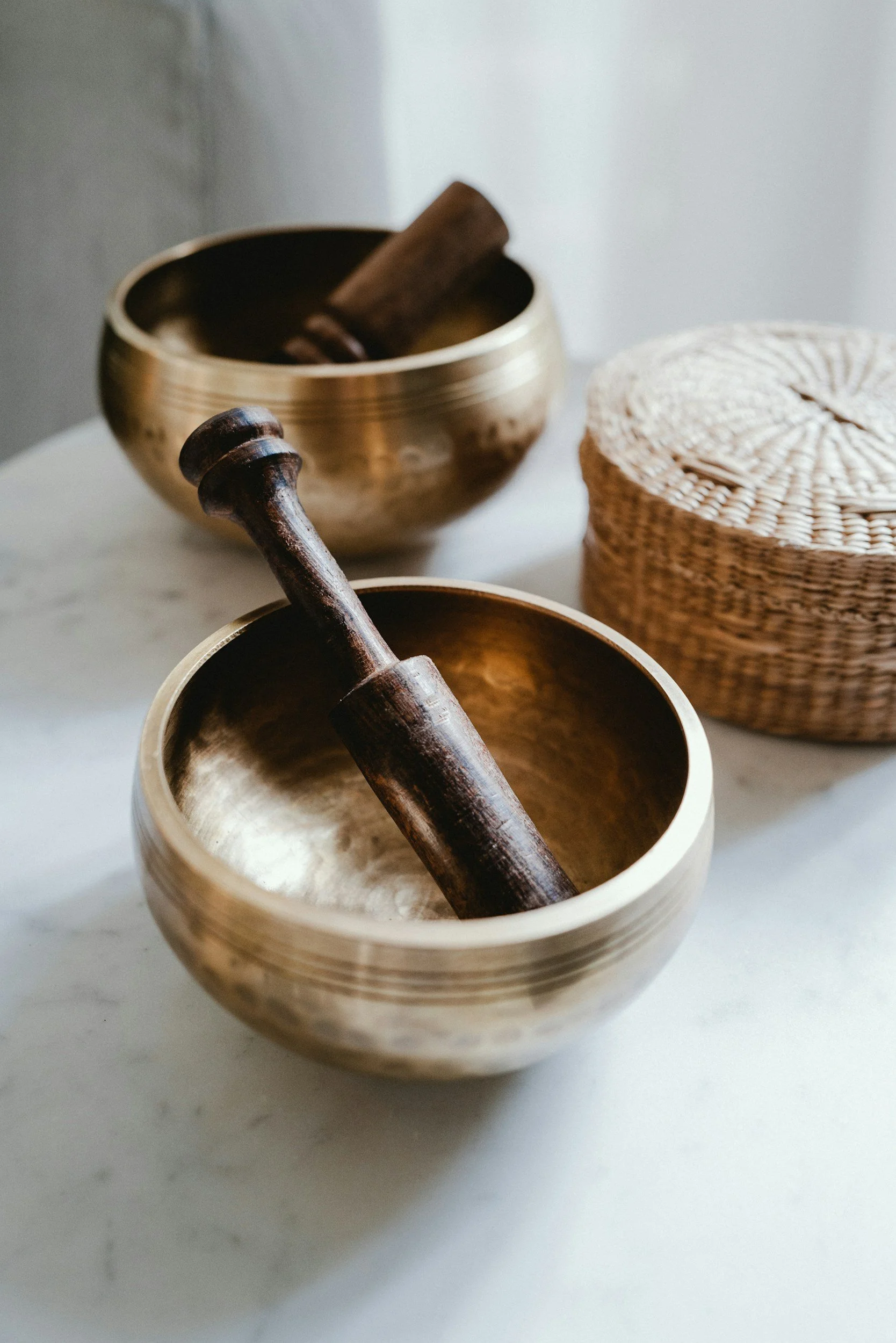 Two brass singing bowls with wooden mallets and a wicker container on a white surface.