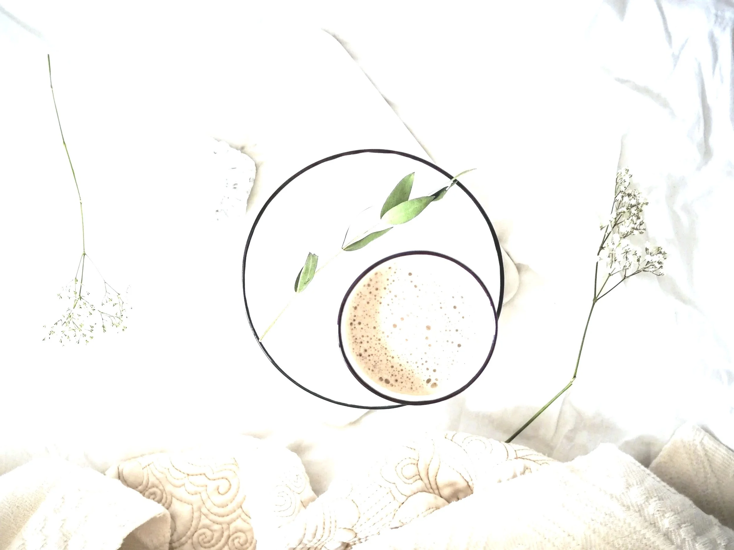 A top view of a white surface with black-rimmed glass cups, one with a creamy beverage and foam, surrounded by delicate white flowers and textured white cloth.