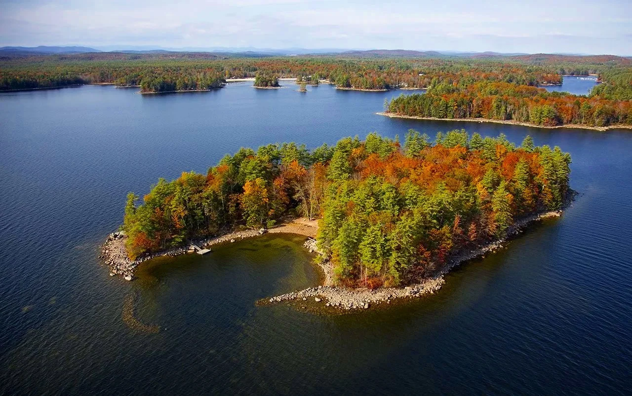 An aerial view of a small, tree-covered island with colorful fall foliage, surrounded by a large lake with other distant islands and forested areas.