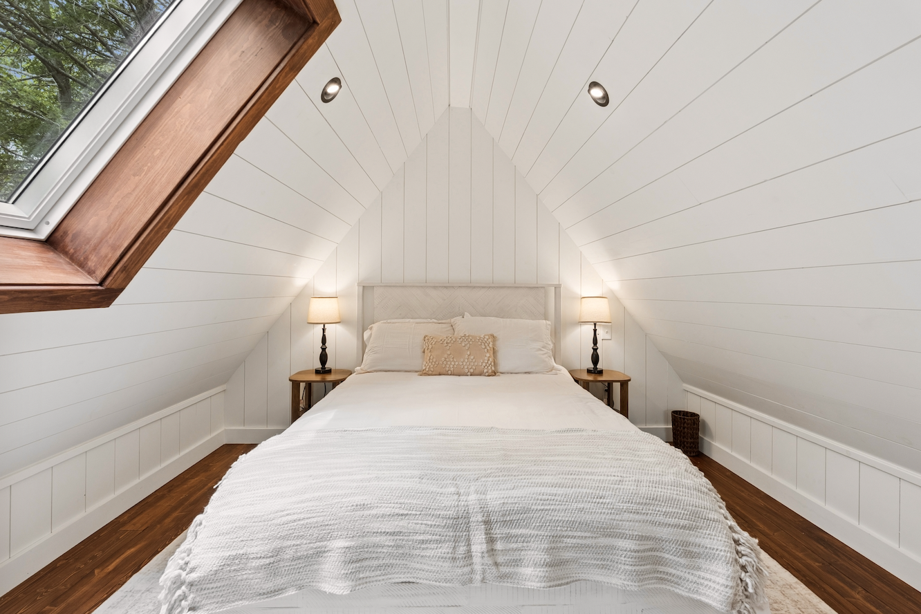 Minimalist bedroom with a sloped ceiling, beige upholstered bed with white bedding, two round bedside lamps, and wooden floors.