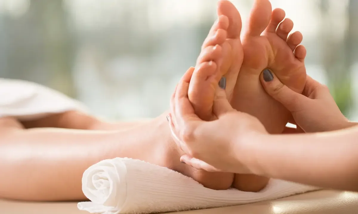 A person receiving a foot massage or reflexology treatment, lying on a massage table with a rolled towel under their ankle, and a therapist using their hands to massage the foot.