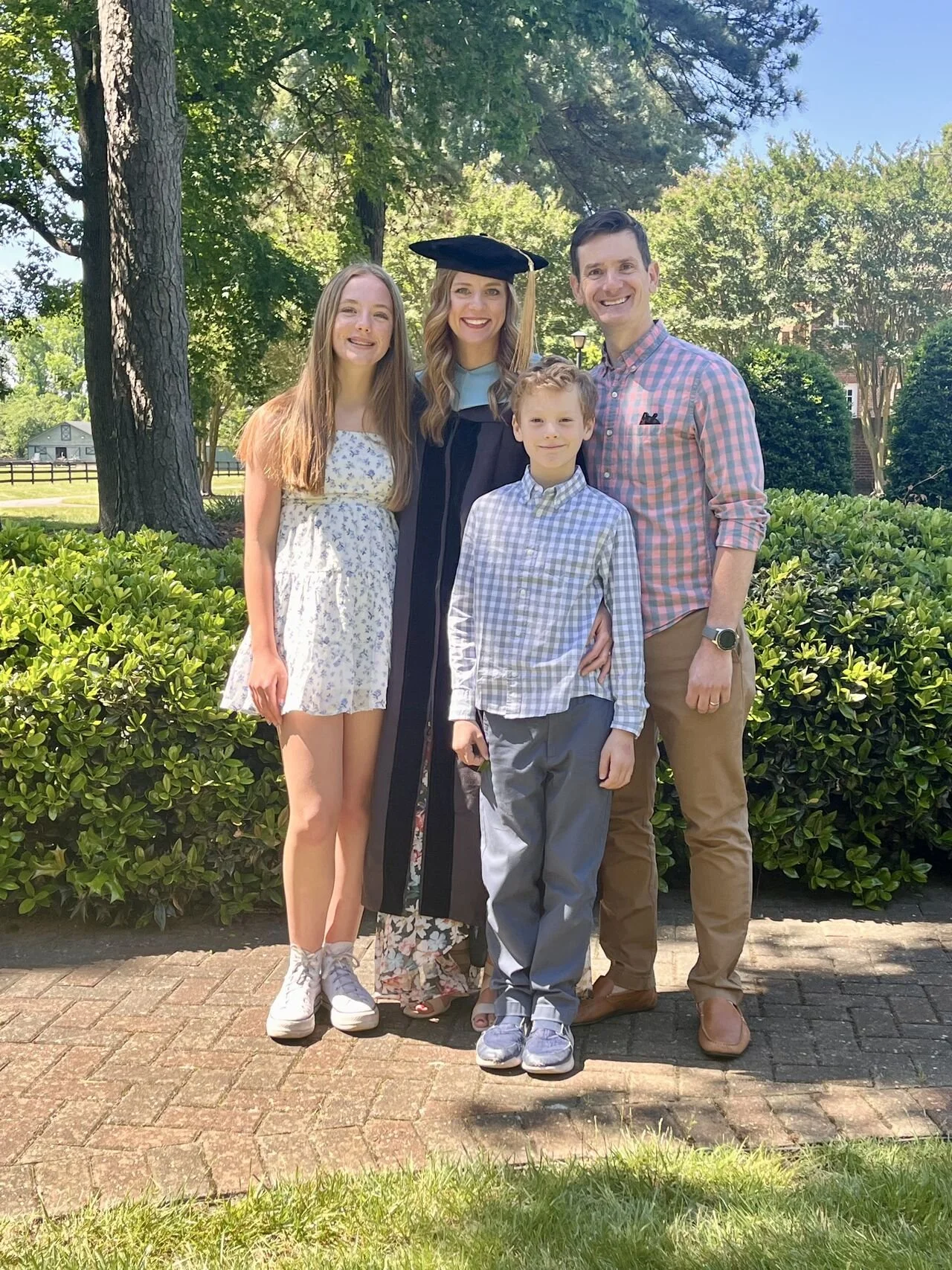 Family celebrating graduation outdoors, with a young woman in cap and gown, posing with a man, a girl, and a boy, all smiling, standing on a brick path surrounded by greenery and trees.