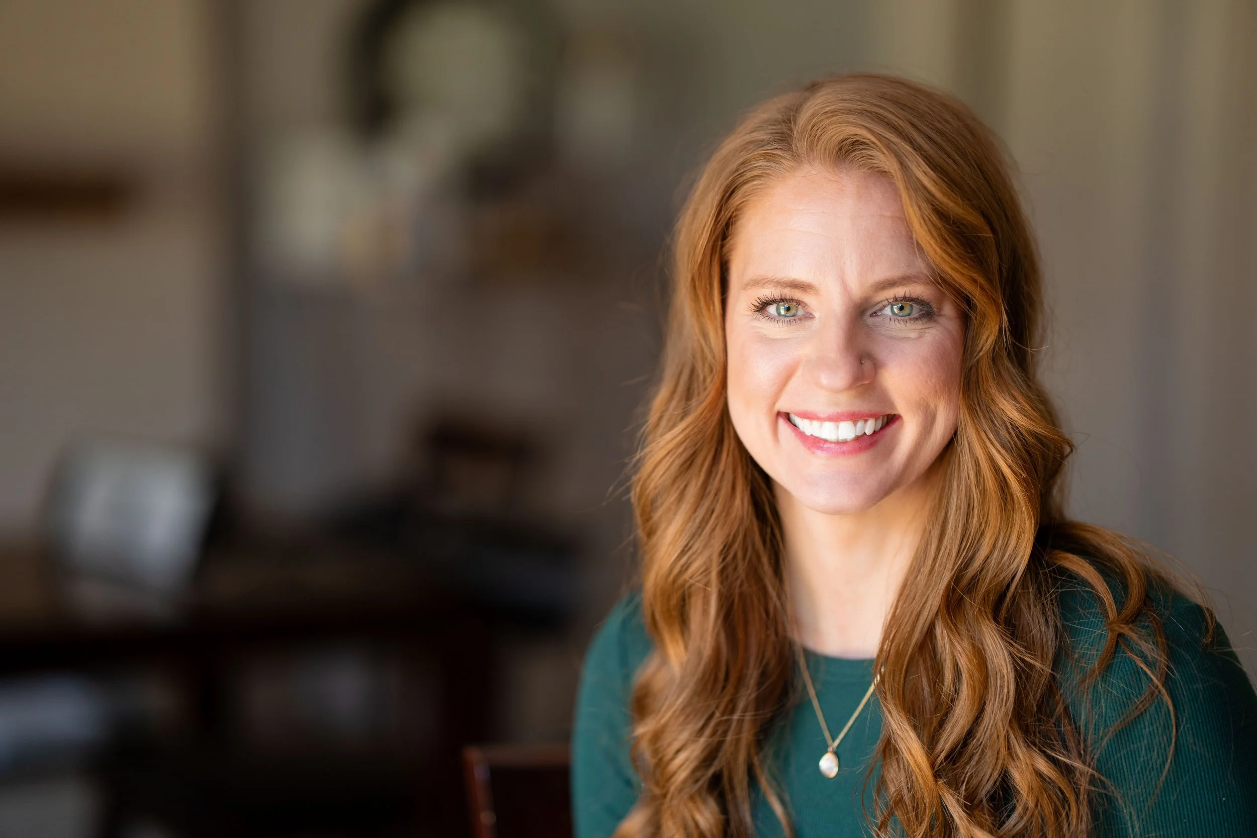 A woman with long, wavy red hair smiling in a room with blurred background.