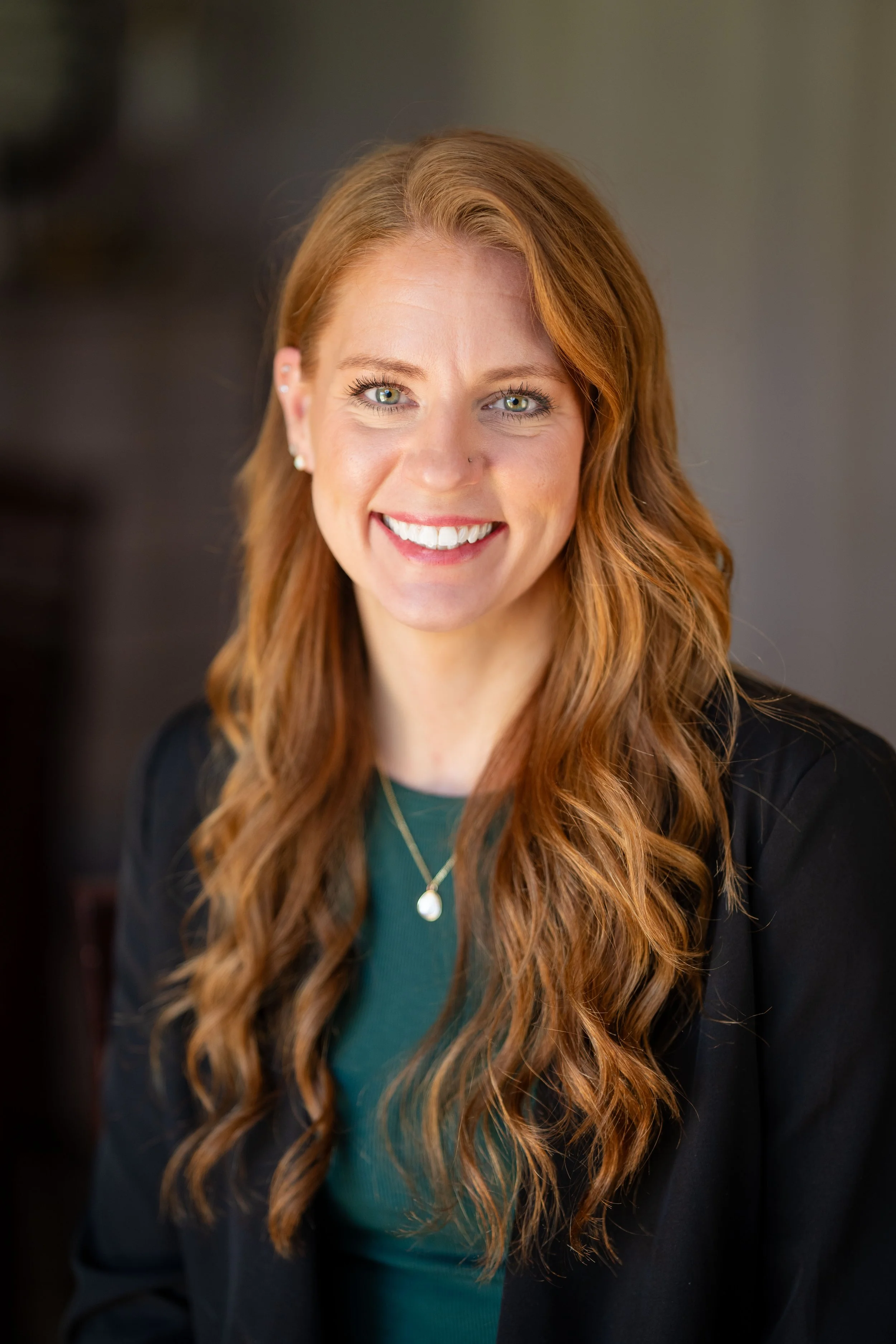 A woman with long wavy red hair, blue eyes, and fair skin is smiling at the camera. She is wearing a dark blazer over a green top, along with a gold necklace with a pendant and pearl earrings.