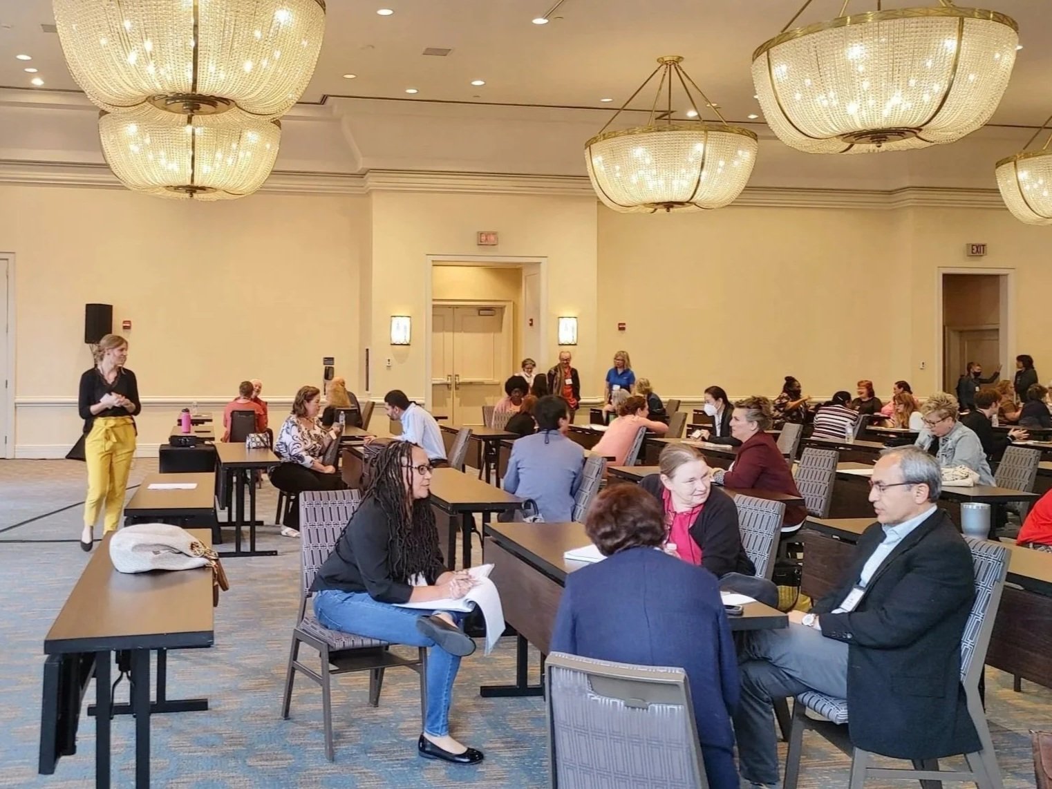 People seated at tables in a large conference room with chandeliers, engaging in discussions and using laptops.