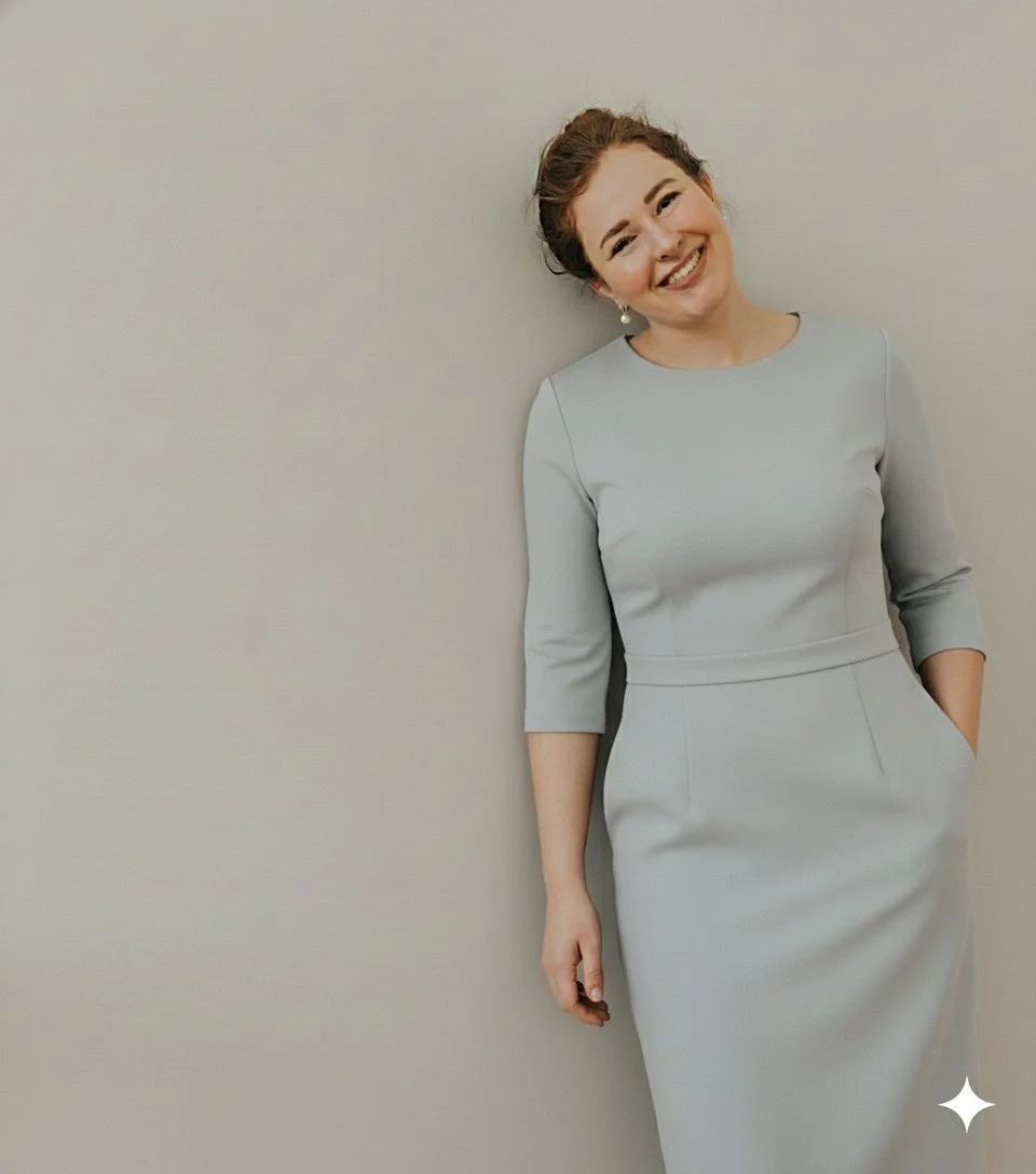 A woman with reddish hair tied back, smiling, wearing a light gray dress with three-quarter sleeves, standing against a neutral wall.