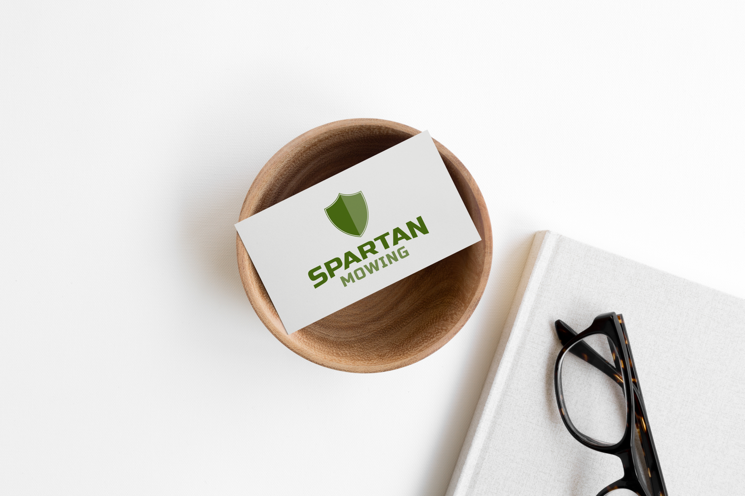 A wooden bowl with a business card that reads "Spartan Mowing" and features a green shield logo, placed on a white desk next to a white book and a pair of black glasses.