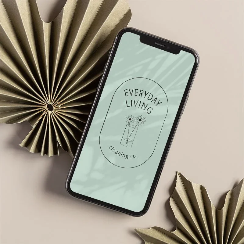 Smartphone displaying a logo for 'Everyday Living Cleaning Co.' with a minimalist flower illustration, placed on a beige surface with decorative gold paper fans.