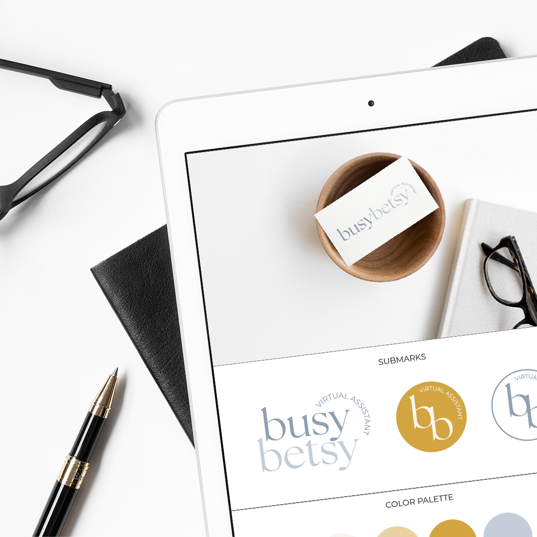 Top-down view of a white desk with a tablet displaying logo options for 'busy betsy' virtual assistant, a black leather notebook, a gold and black pen, glasses, and a small wooden bowl with a business card reading 'busybet sy' inside.