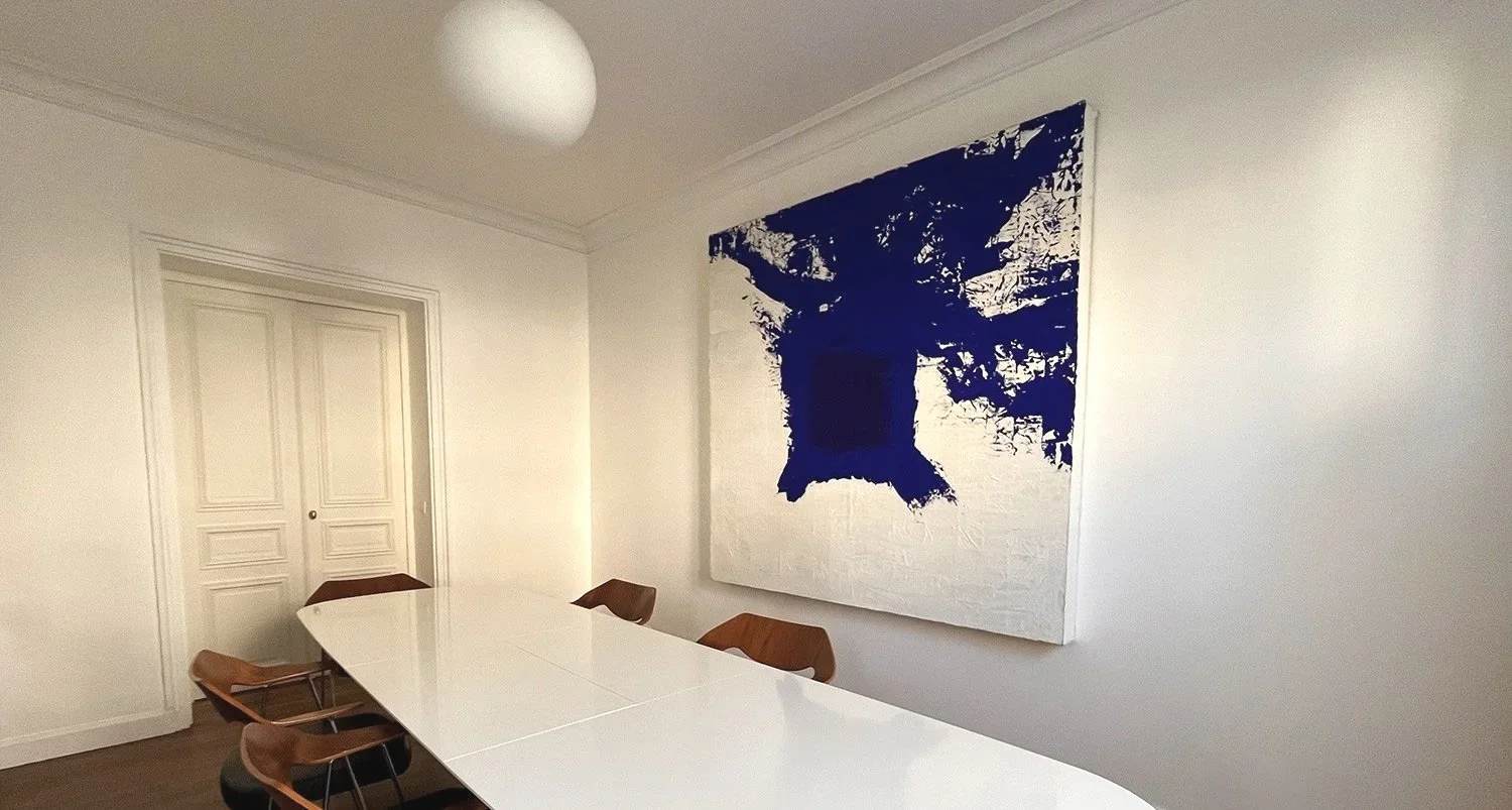 Salle de réunion simple avec une grande table blanche, six chaises en bois, un tableau abstrait bleu et blanc sur le mur, et une lumière suspendue au plafond.