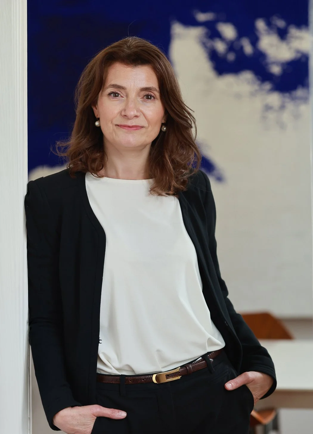 Femme d'affaires avec des cheveux bruns, portant un tailleur noir et une chemise blanche, posant dans un bureau avec un tableau du monde en arrière-plan.