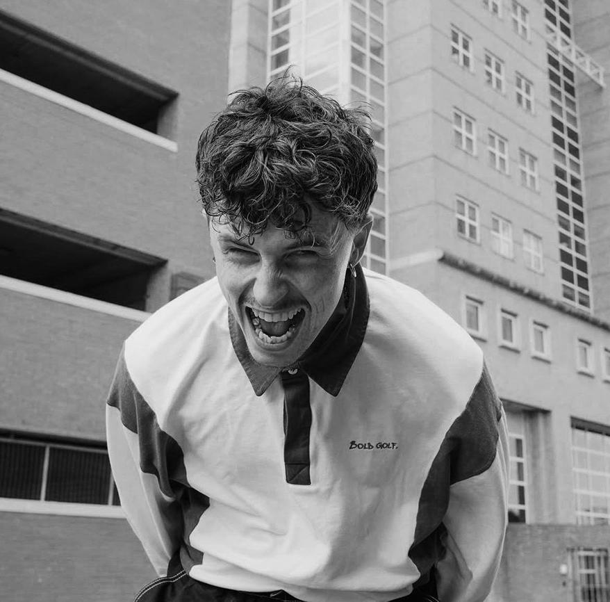 Young man with curly hair laughing outdoors in front of buildings.