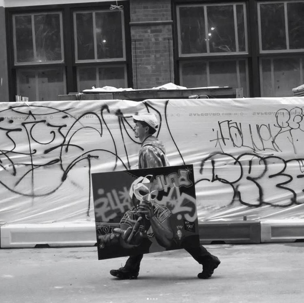 A person walking on the street carrying a large spray-painted picture of a young man wearing a cap and holding a spray can. Behind, a construction site with graffiti-covered barriers and a building with large windows is visible.