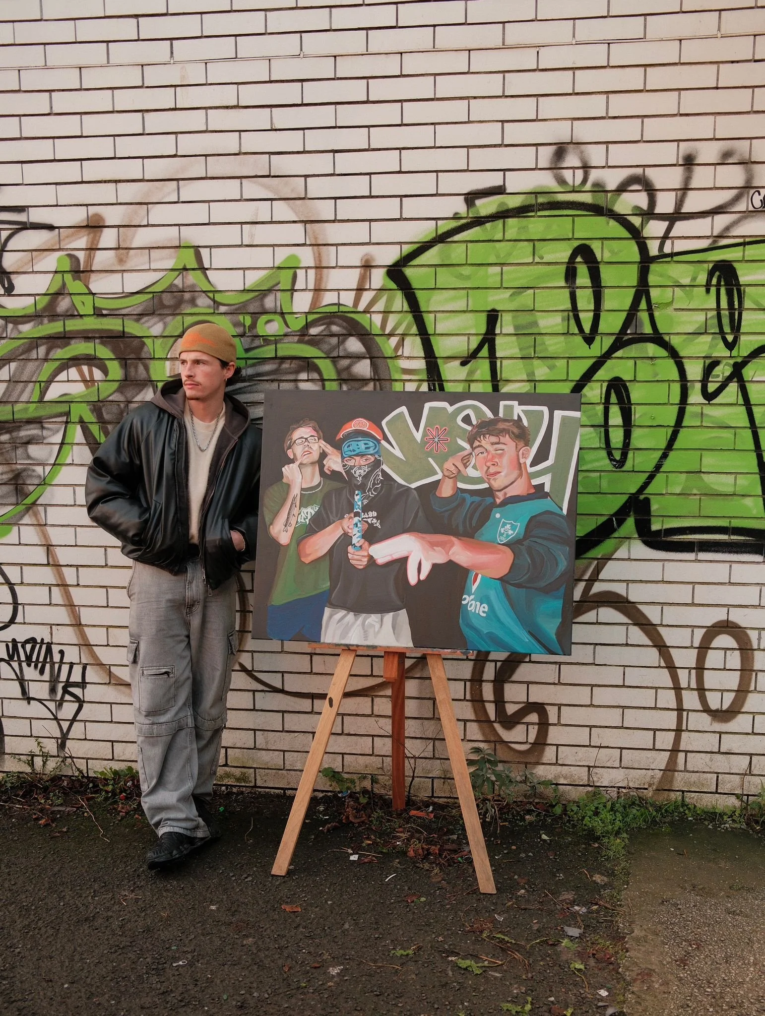 A young man with a mustache, wearing a tan beanie, black jacket, grey sweatpants, and black shoes, stands by a graffiti-covered brick wall while leaning against a large art poster on an easel. The poster depicts three young men, one with glasses and tattoos, the other wearing a mask, and the third making a gesture with his hand.