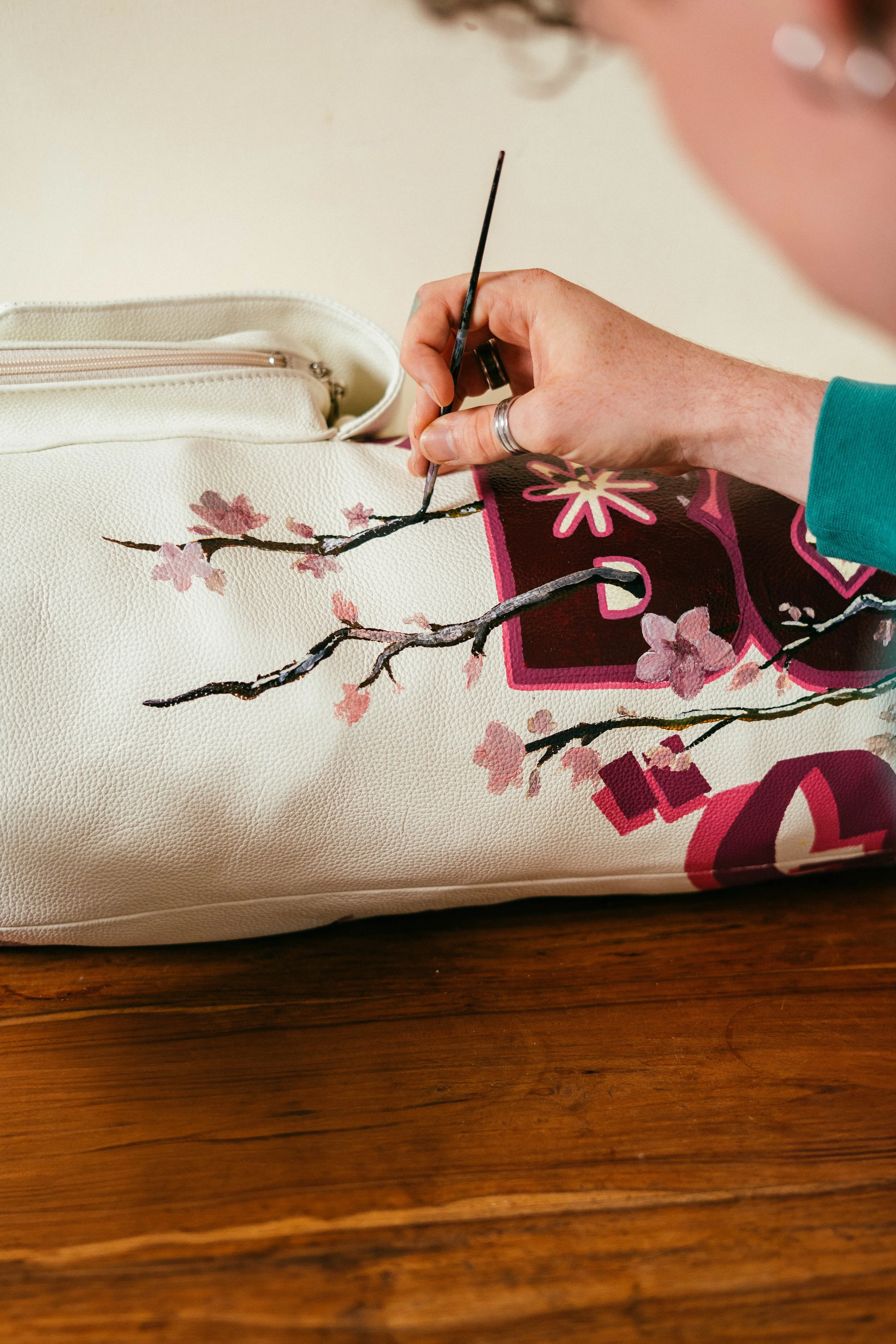 Close-up of a person's hand painting pink cherry blossoms on a cream-colored surface, with a beige zippered pouch nearby.