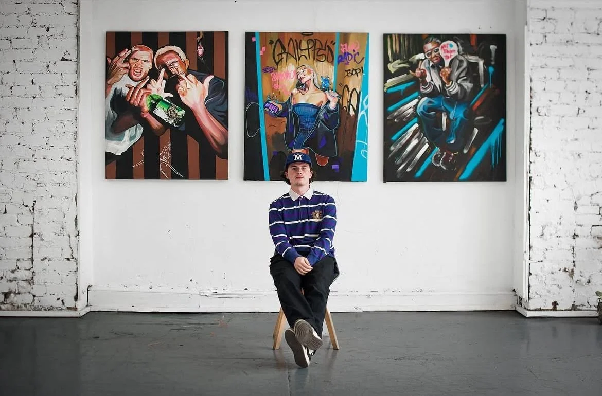 A young man sitting on a stool in an art gallery, with three graffiti-style paintings on the wall behind him.