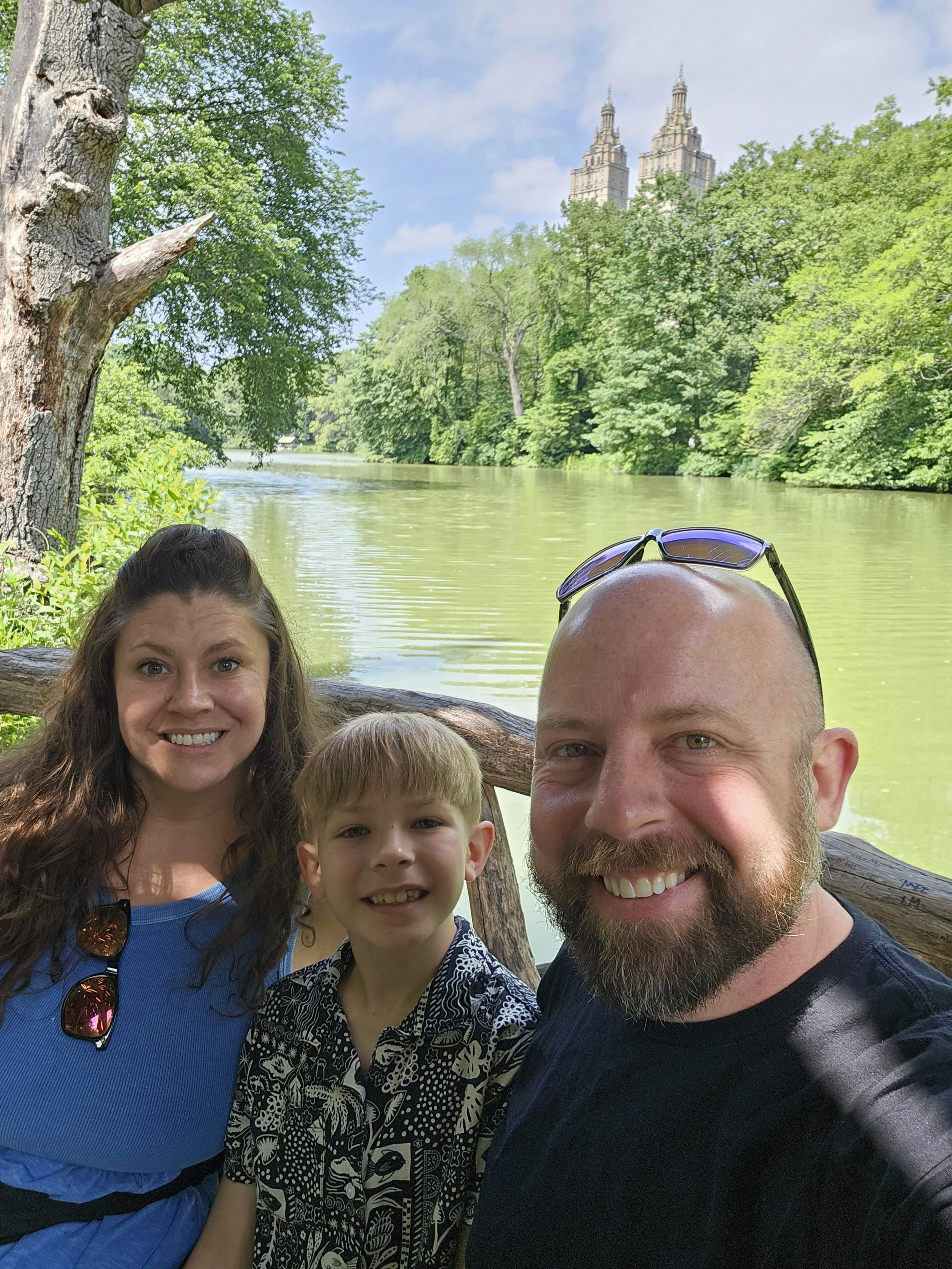 A family of three taking a selfie outdoors by a river with many trees and a city skyline with tall buildings in the background.