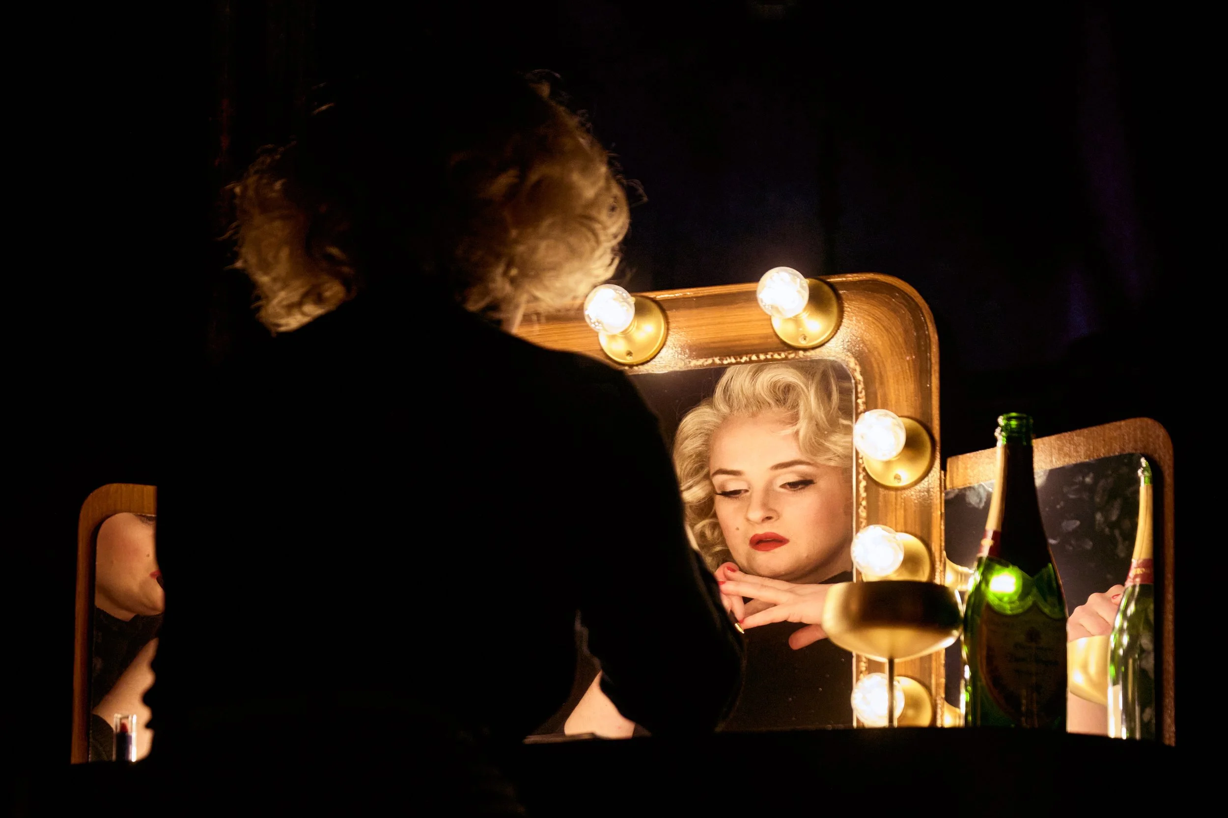 Marilyn Monroe (Veronika Schäfer) schaut in einen beleuchteten Schminkspiegel, Getränke auf dem Tisch, dunkler Hintergrund. ©Joseph Ruben Heicks