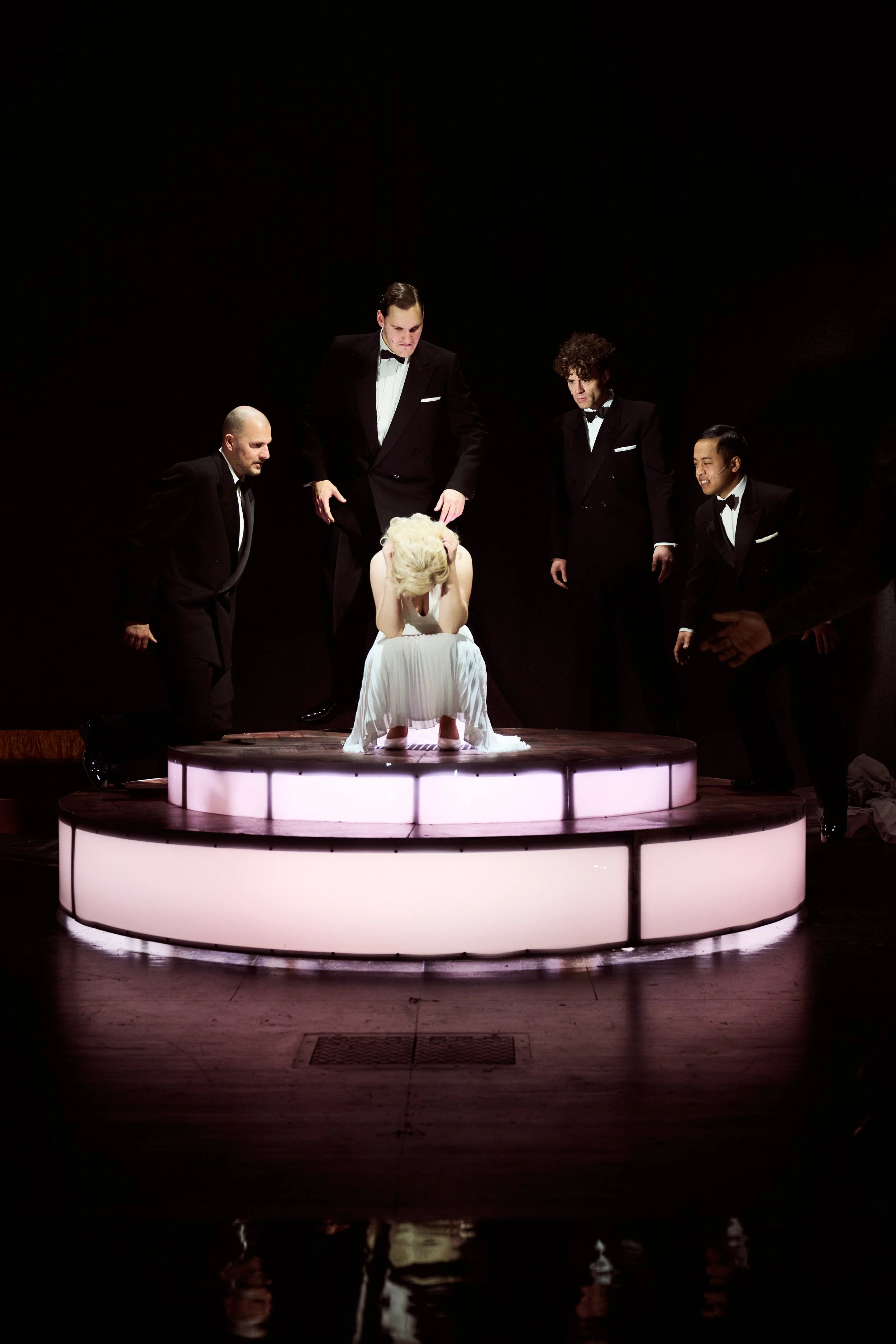 Marilyn Monroe (Veronika Schäfer), die auf Knien sitzt und ihren Kopf in den Händen hat, umgeben von fünf Männern (The Tritones) in schwarzen Anzügen auf einer runden Bühne. ©Joseph Ruben Heicks