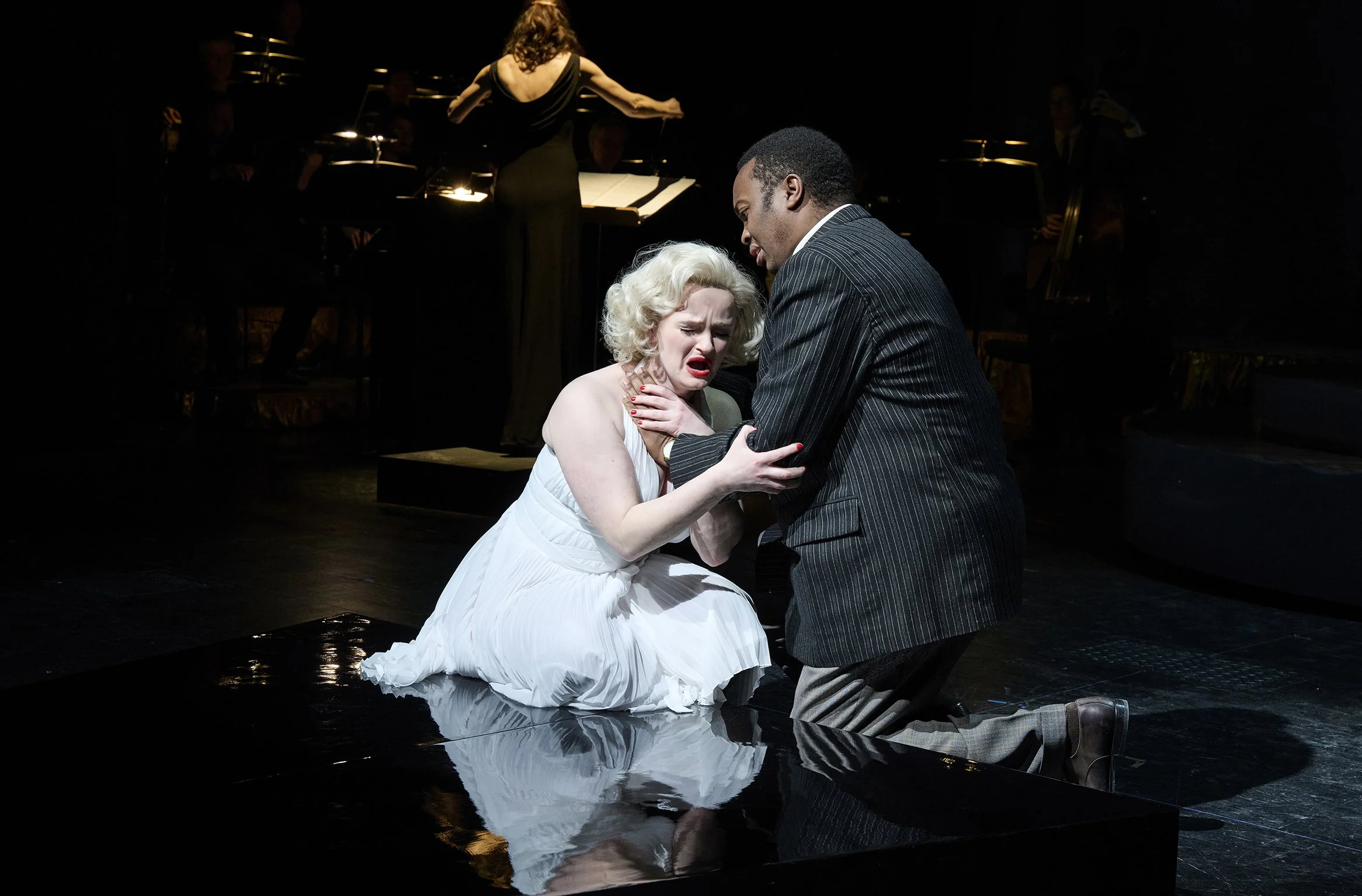 Marilyn Monroe (Veronika Schäfer) in einem weißen Kleid, die auf den Knien auf einer schwarzen Bühne sitzt, und einem Mann (Zaccharia Kariithi) in einem dunklen Anzug, der sie berührt. ©Thomas M. Jauk