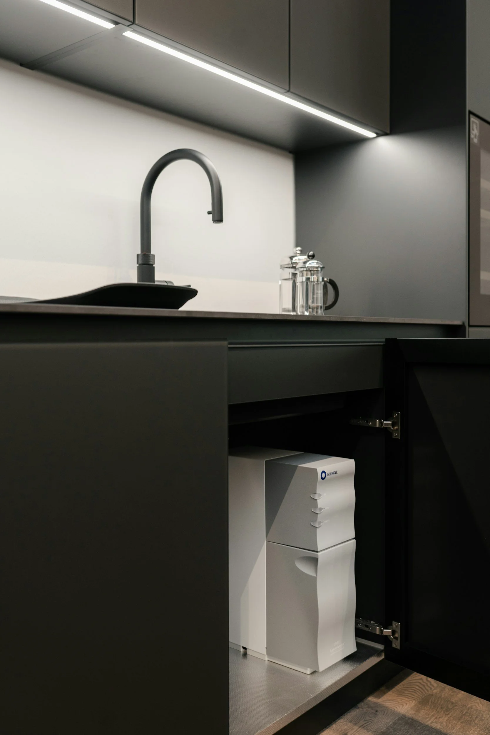 Moderne schwarze Küchenschränke mit offenem Fach, darin ein Luftreiniger, schwarzes Spülbecken mit Wasserhahn, auf der Arbeitsplatte Glasbehälter, dunkle Wand und lichter Schatten