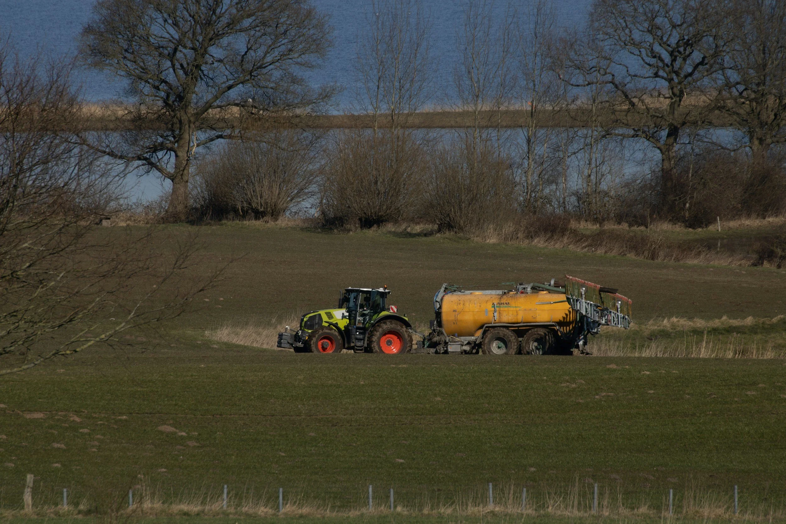 Landwirtschaftliches Fahrzeug mit Gelb-Tank auf grünem Feld, Bäume im Hintergrund, See und Himmel im Abstand.