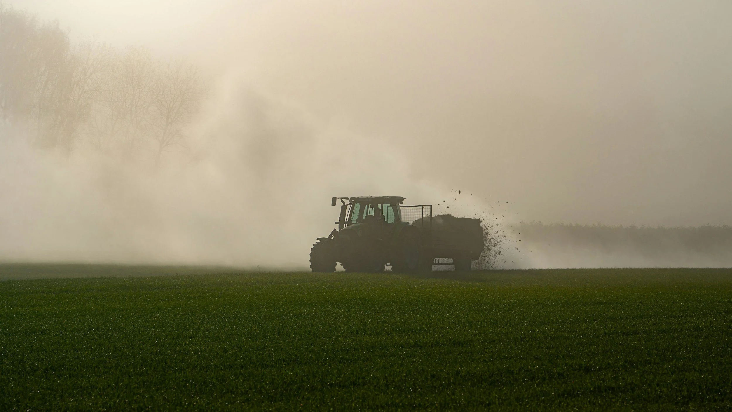 Traktor bei der Arbeit auf einem Feld, Staub und Rauch in der Luft, bewölkter Himmel im Hintergrund.