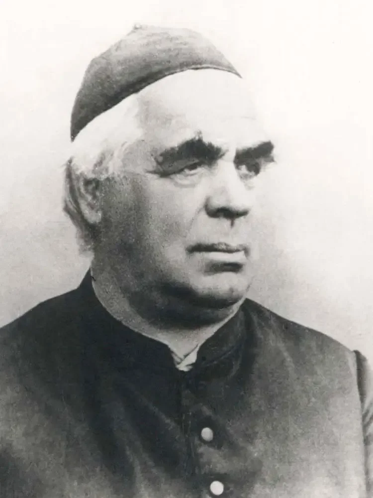 Schwarz-weiß Foto eines Mannes mit traditioneller Kleidung und einer Mütze, in Nahaufnahme, ernster Gesichtsausdruck.