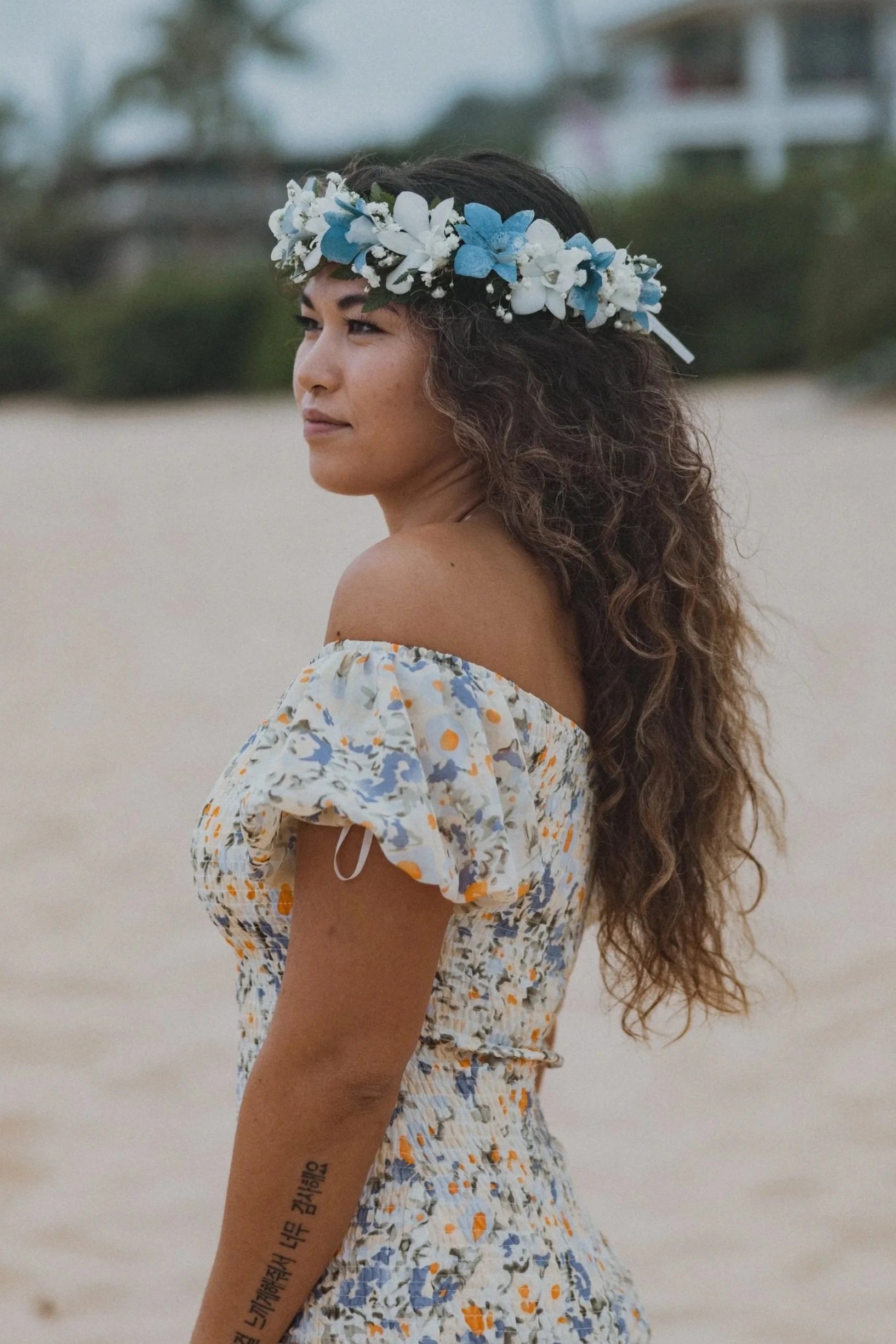 A woman with long, curly hair wearing a floral dress and a white and blue flower crown, standing on a beach.