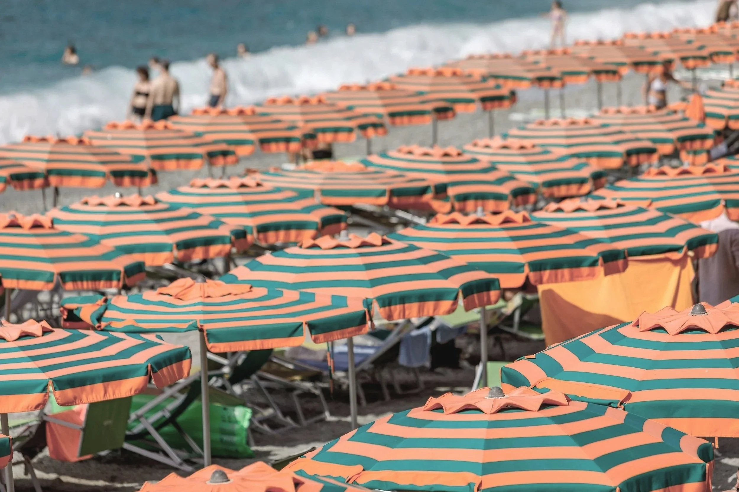 Many orange and green striped beach umbrellas lined up on a sandy beach with lounge chairs underneath, some people standing near the water's edge in the background.