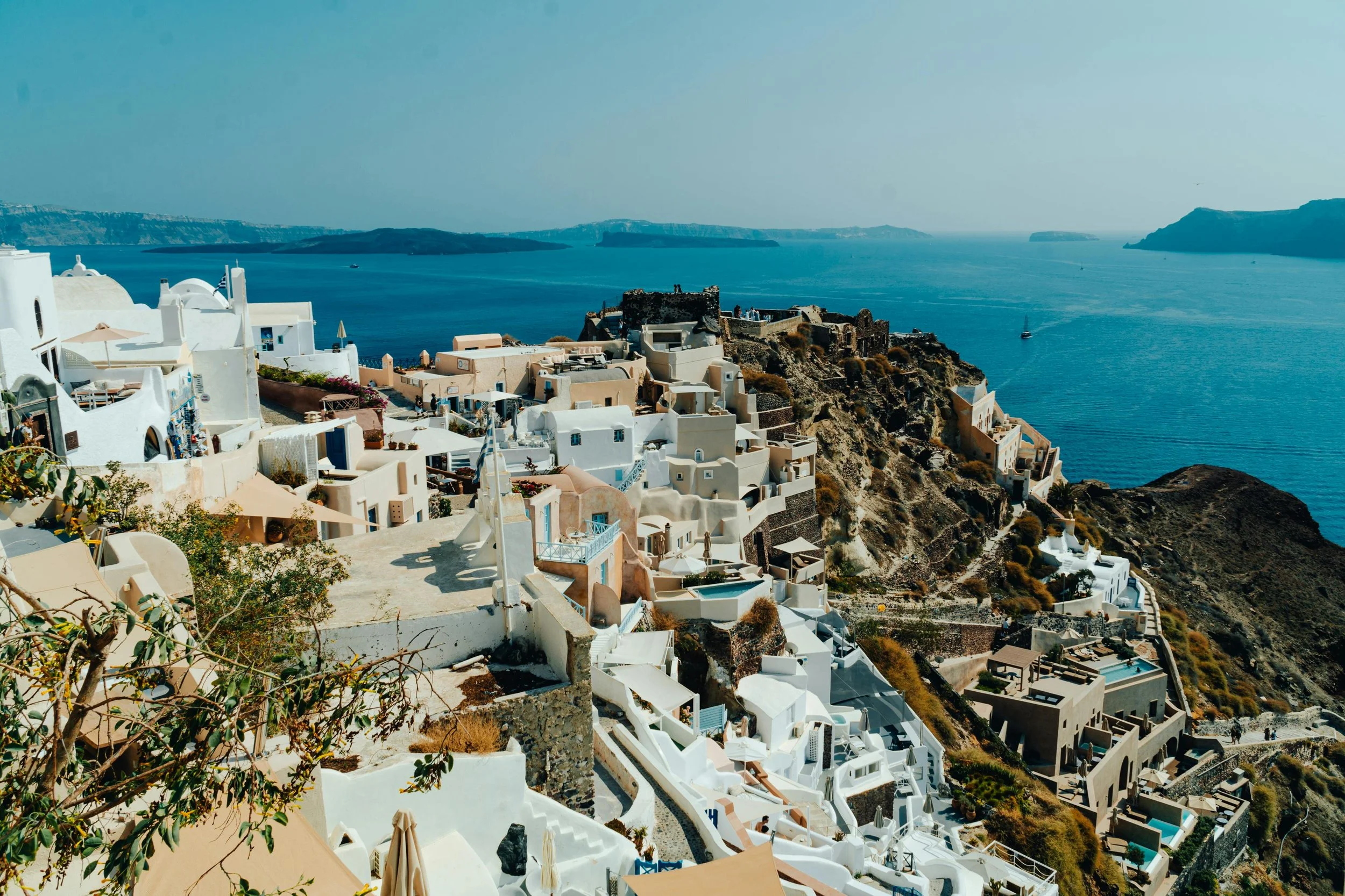 White buildings with blue accents on a steep hillside overlooking the deep blue sea, with a clear sky and some boats in the water.