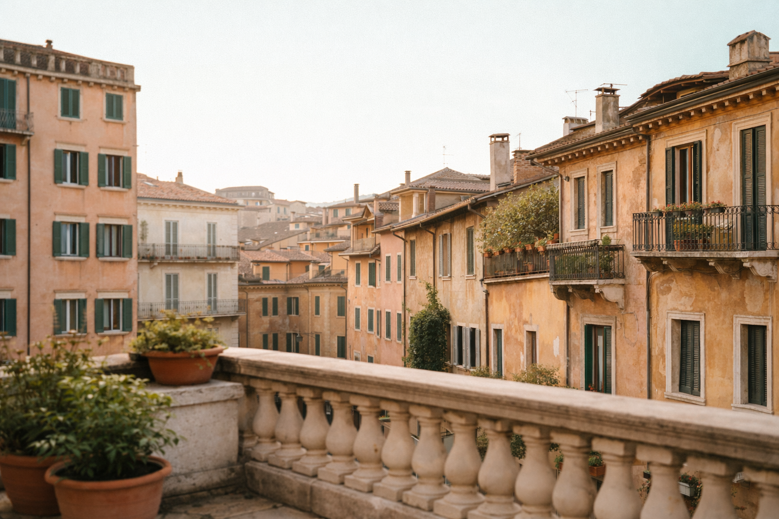 View of old European-style apartment buildings with ochre and beige facades and green shutters, seen from a balcony with potted plants and a stone railing, during daytime.
