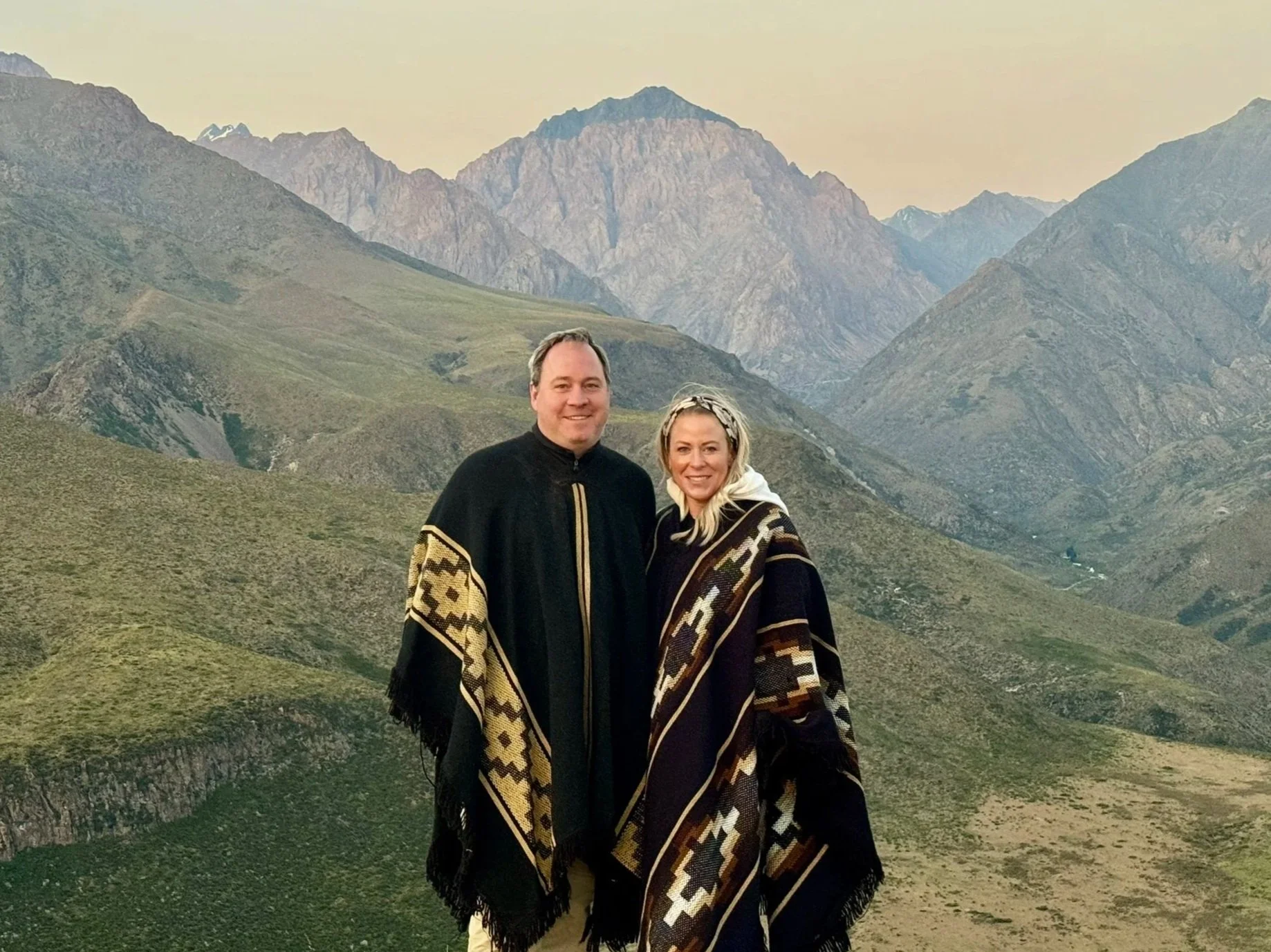 A man and woman standing outdoors with the Andes mountain range in the background, both wearing traditional patterned shawls.